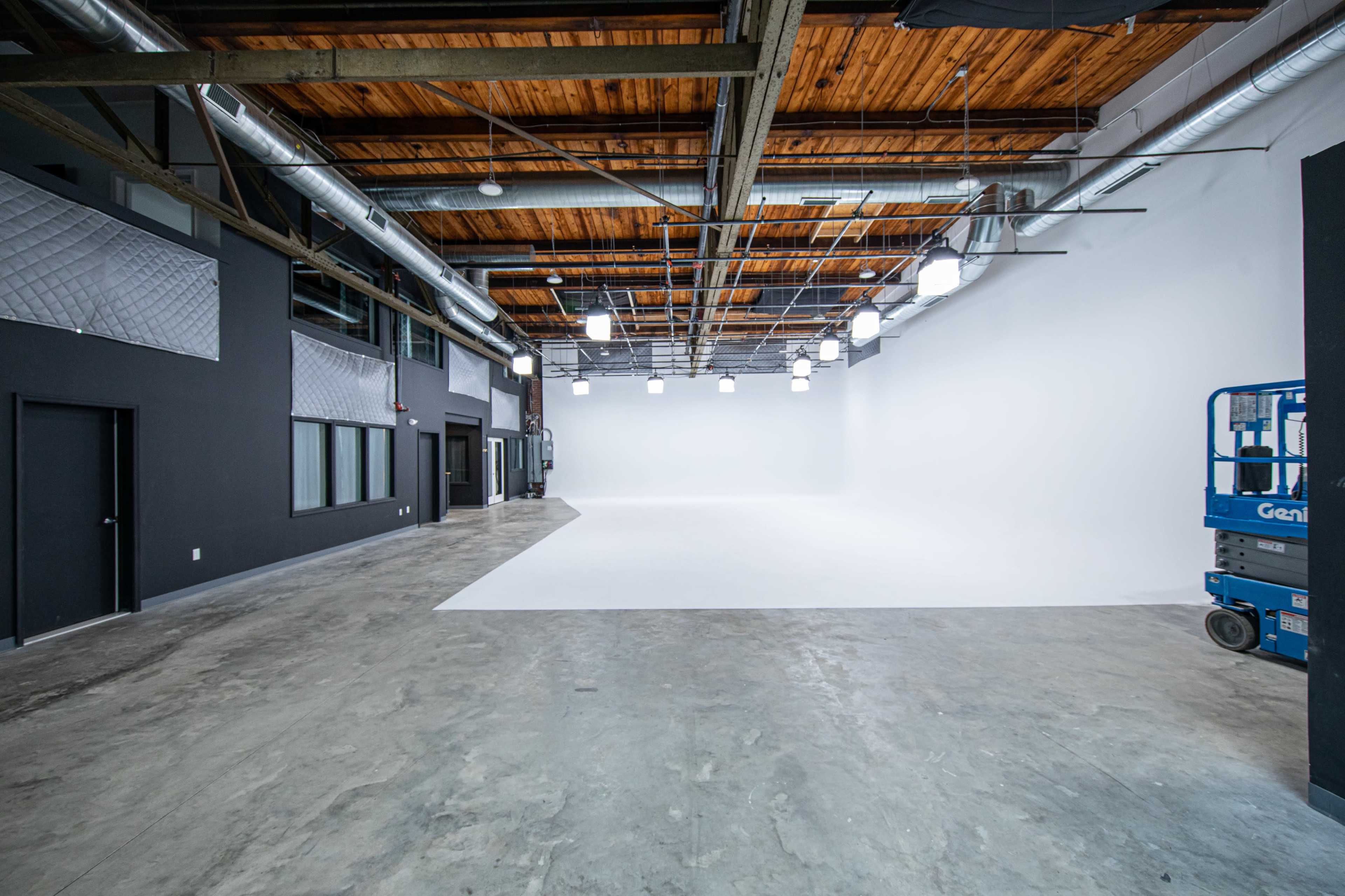 A spacious studio with a white backdrop, concrete floors, exposed wooden beams, and overhead lights.