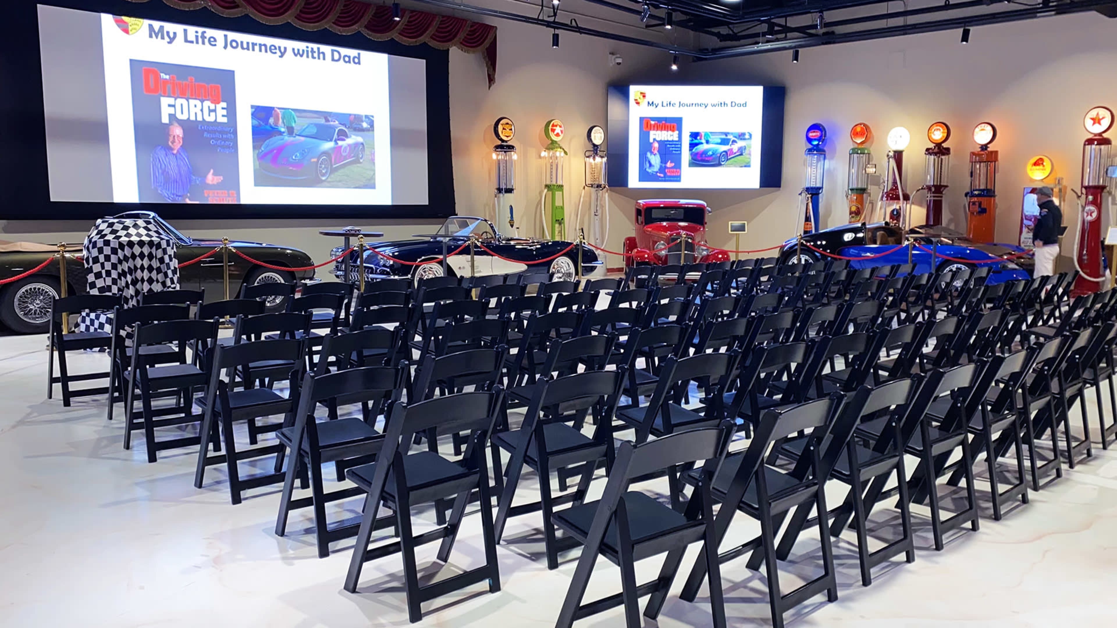 A venue set up for a presentation, featuring rows of black chairs, vintage cars on display, and screens showing slides about a personal journey, along with retro gas station pumps in the background.