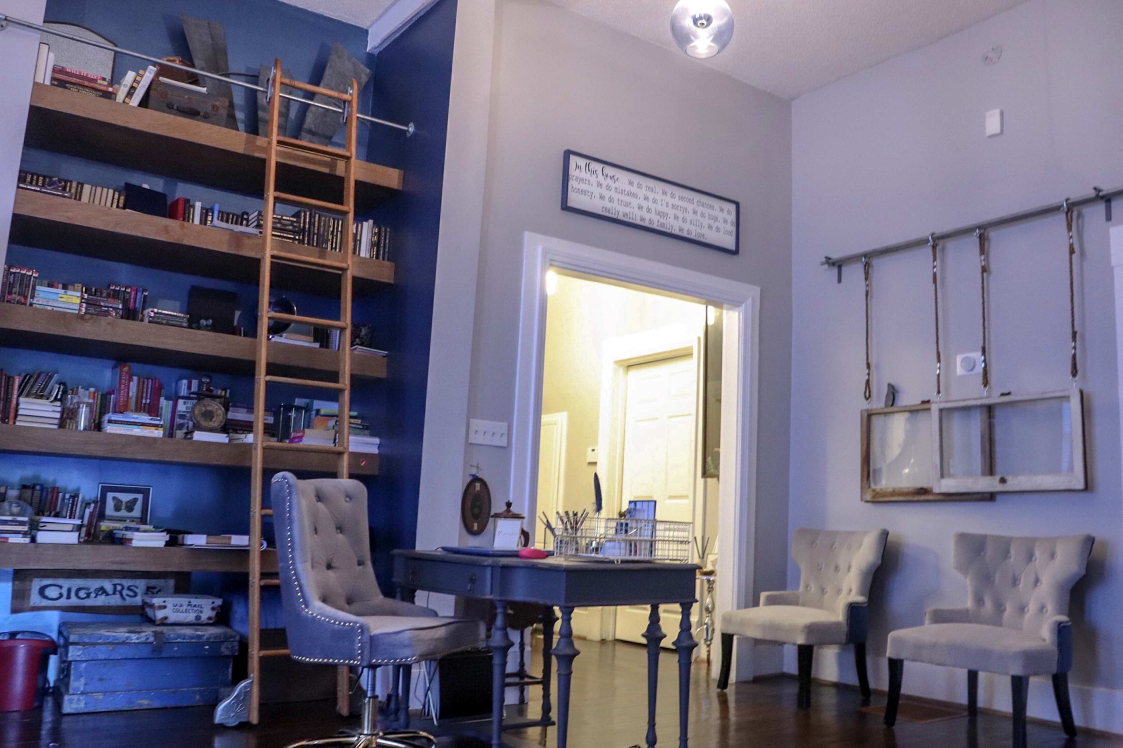 A cozy office space with a wooden bookcase filled with books, a desk with a chair, and three upholstered chairs against a wall.