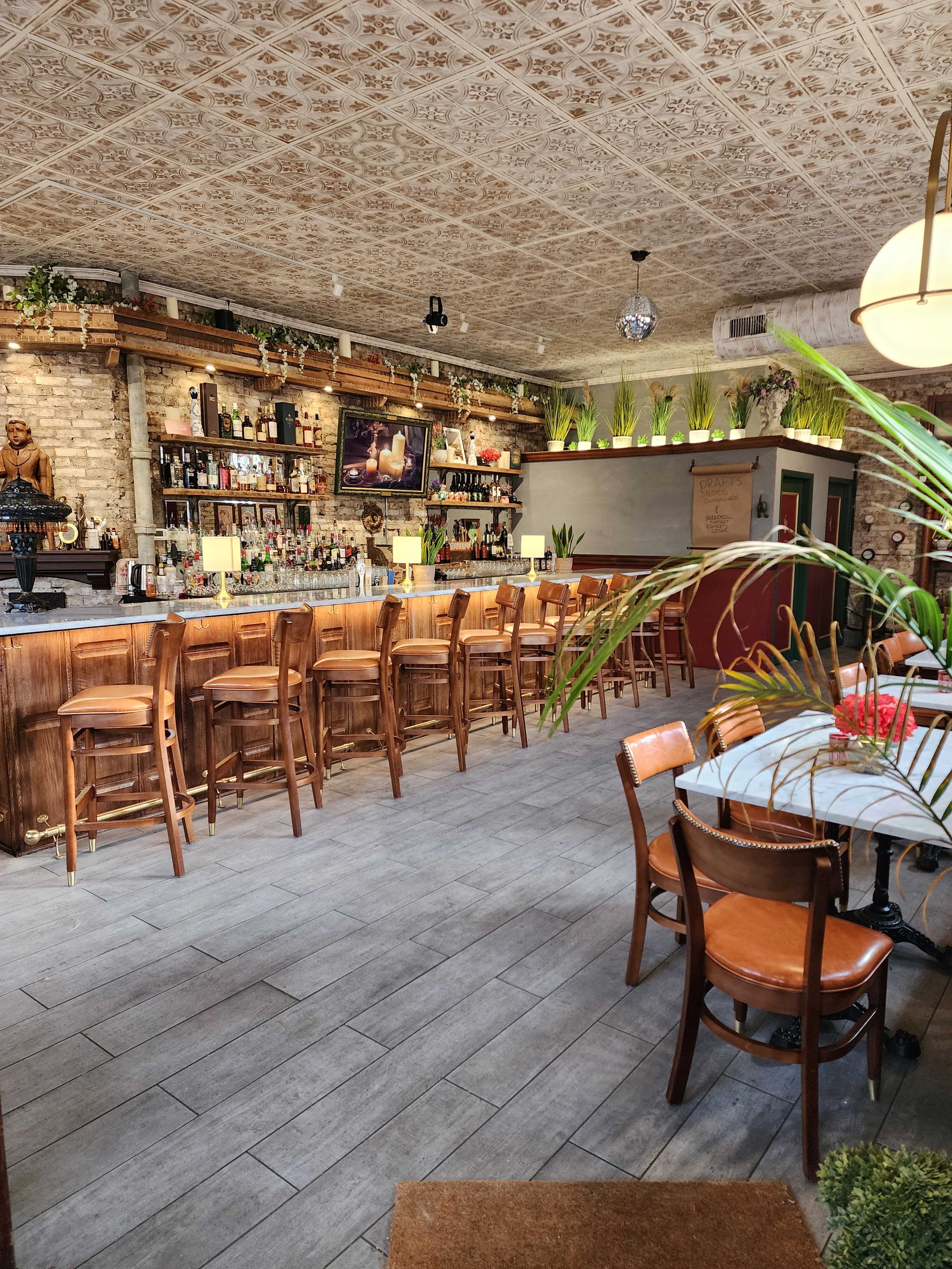 The image shows a bar area with wooden stools and a counter, surrounded by tables, decorative plants, and shelves filled with bottles.