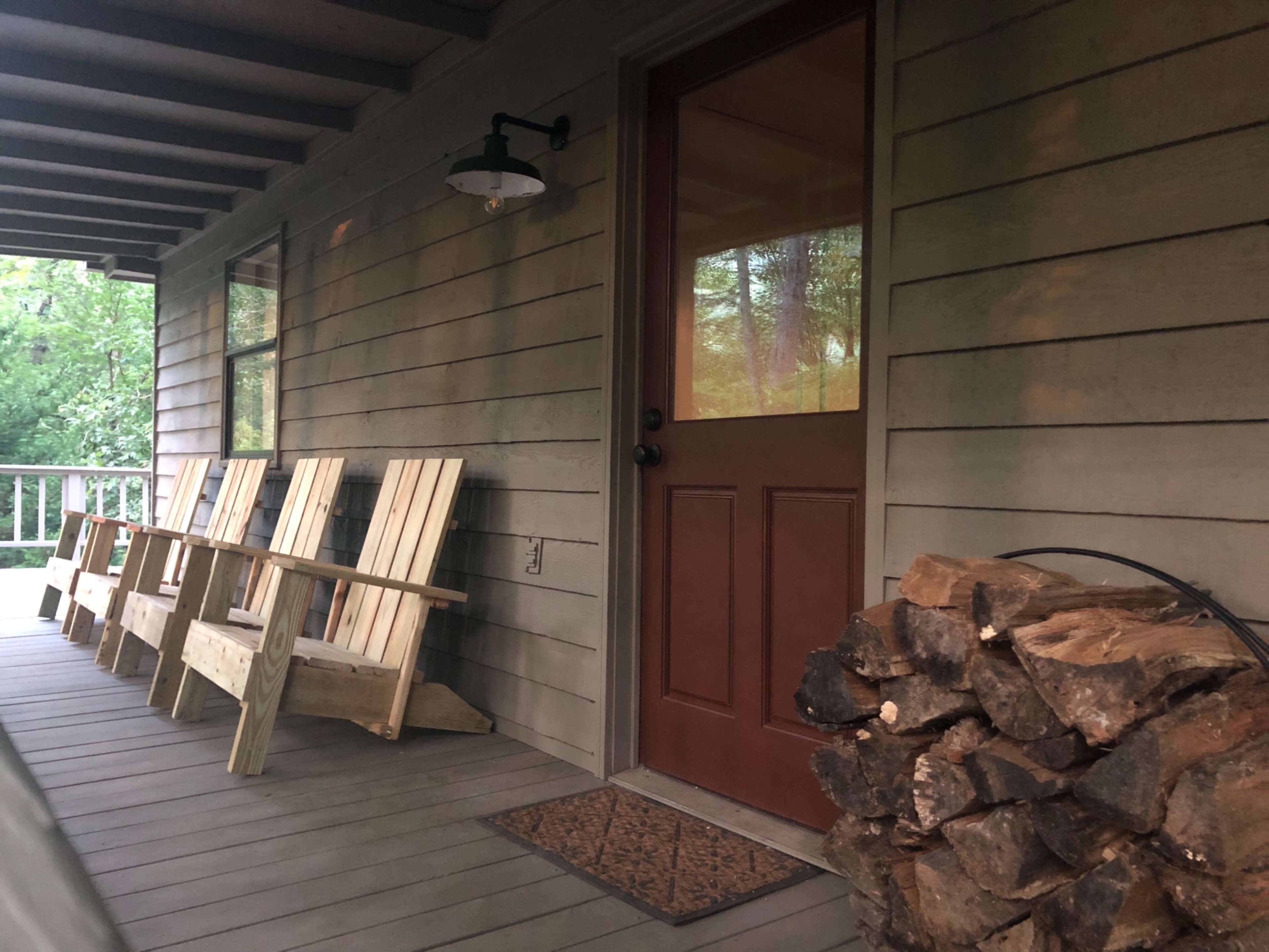 The image shows a porch with several wooden chairs lined up against the wall, a red door, and a stack of firewood beside it.