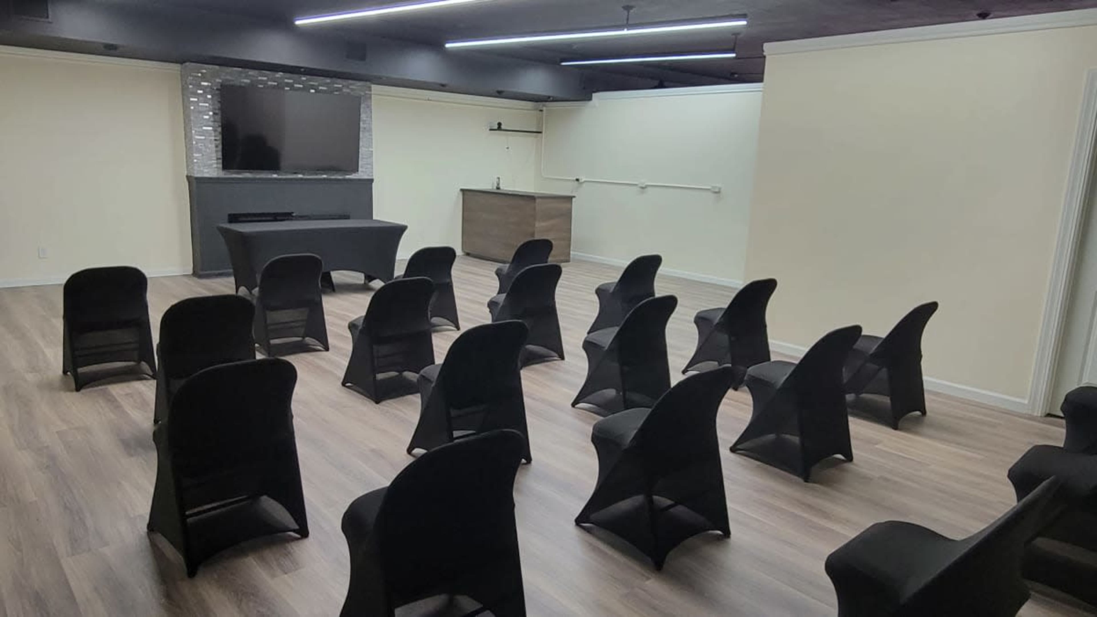 A spacious room set up with rows of black chairs facing a wall-mounted television and a small table, featuring light-colored walls and wooden flooring.