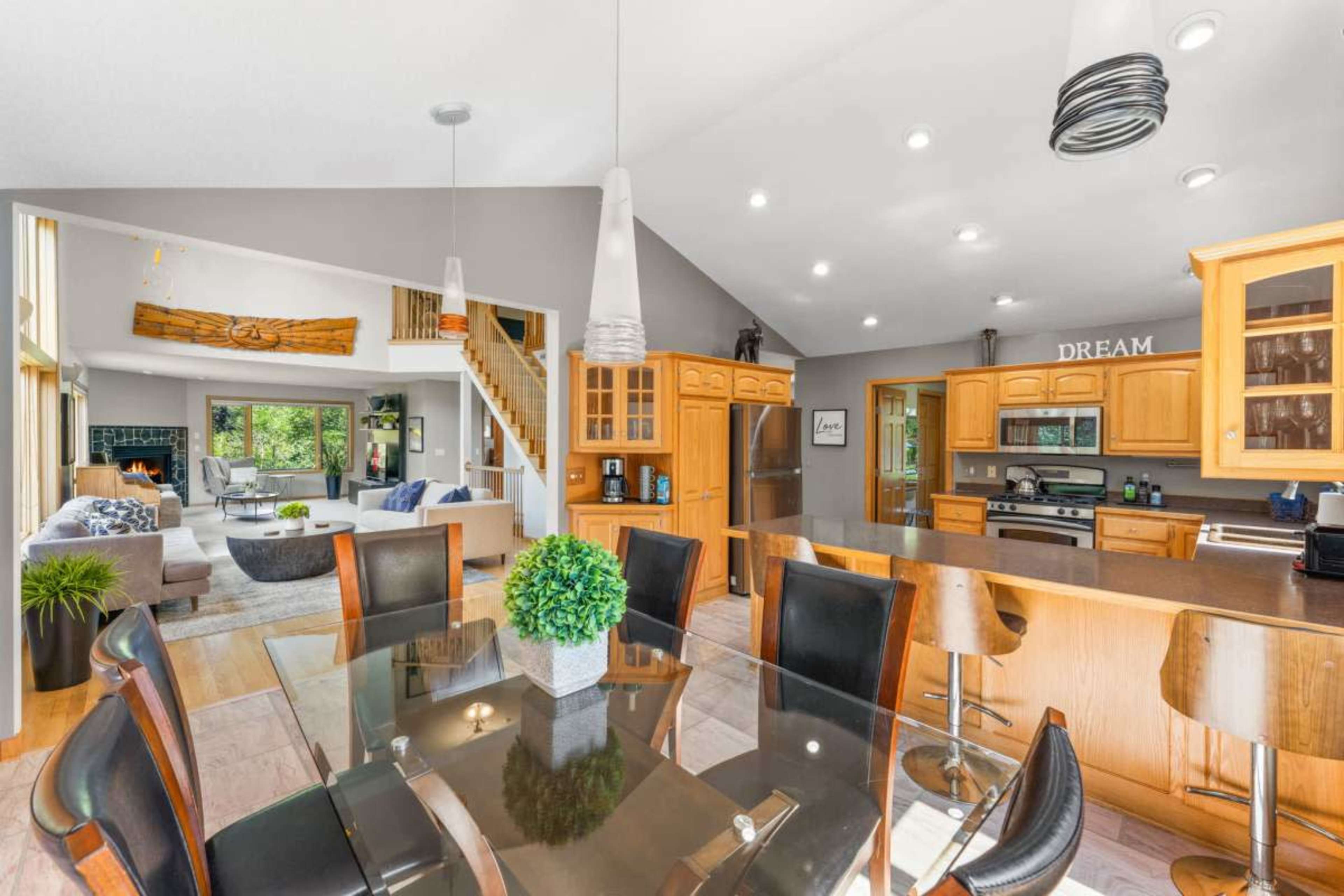 A modern open-concept kitchen and dining area features wooden cabinetry, a glass dining table, and a cozy living space with large windows that allow natural light to fill the room.