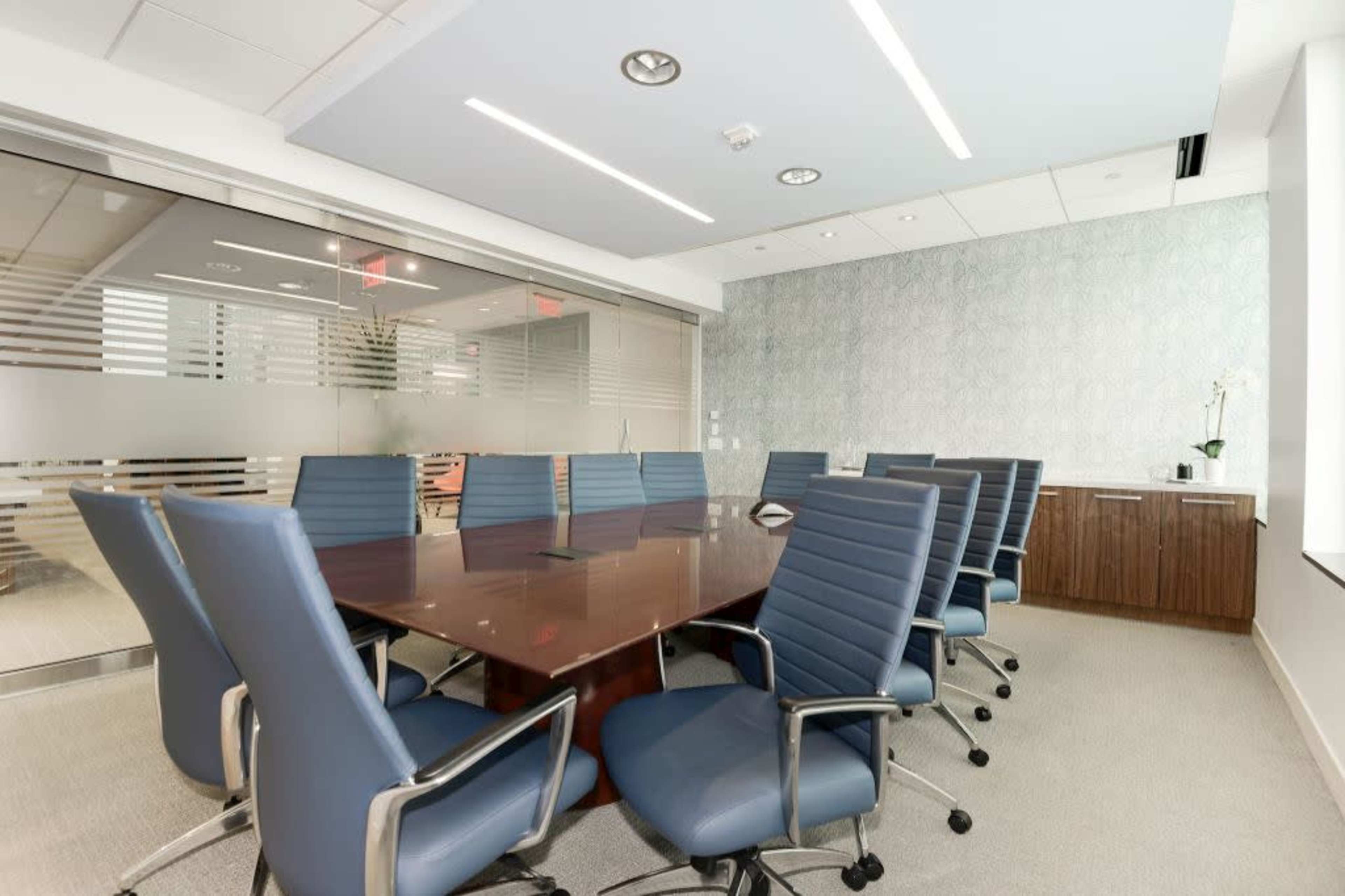 A modern conference room features a large wooden table surrounded by eight blue chairs, with glass walls and a decorative background.