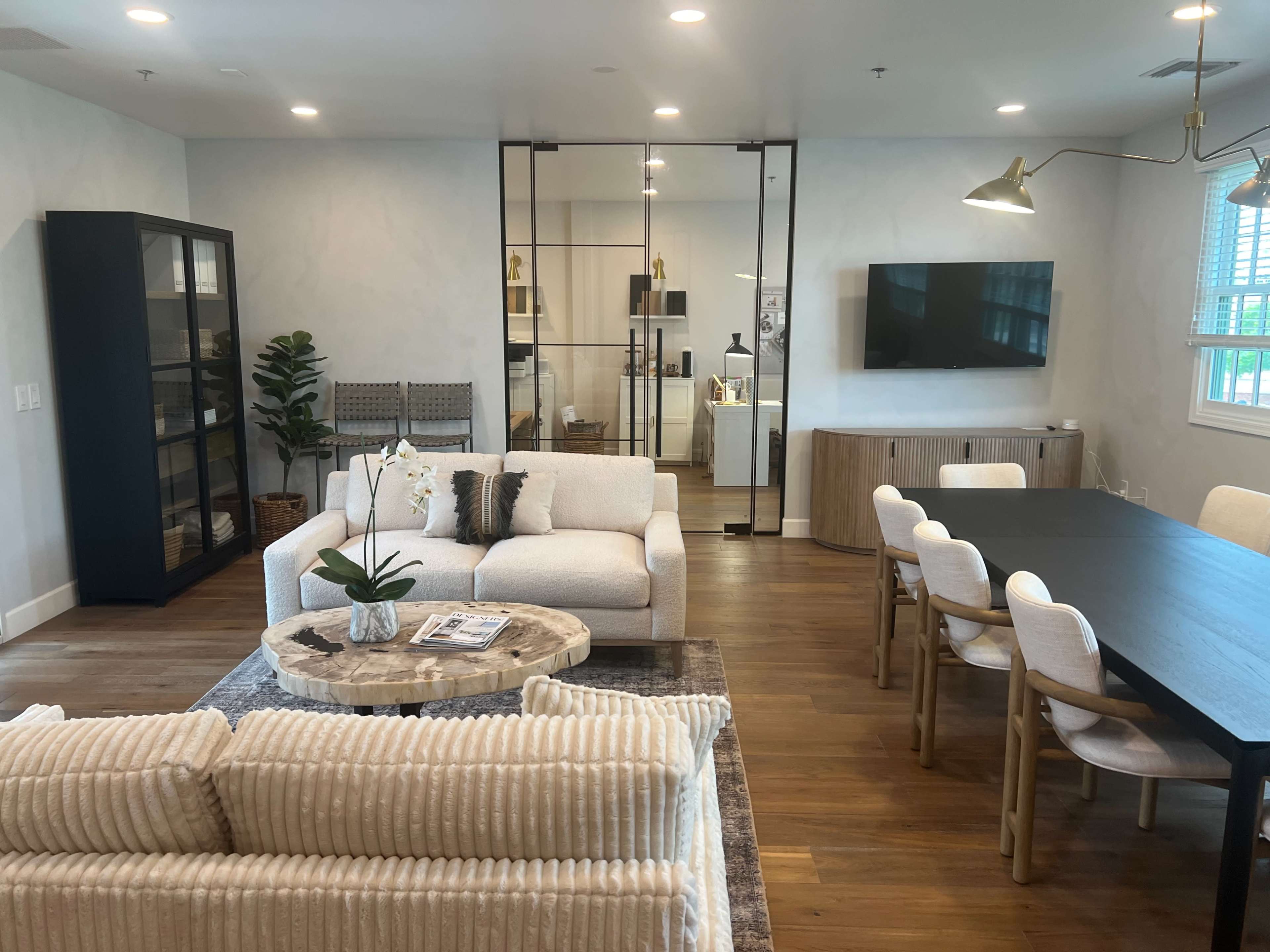 A modern living room and dining area with a sectional sofa, a round coffee table, a dining table, and glass partitions leading to another space.