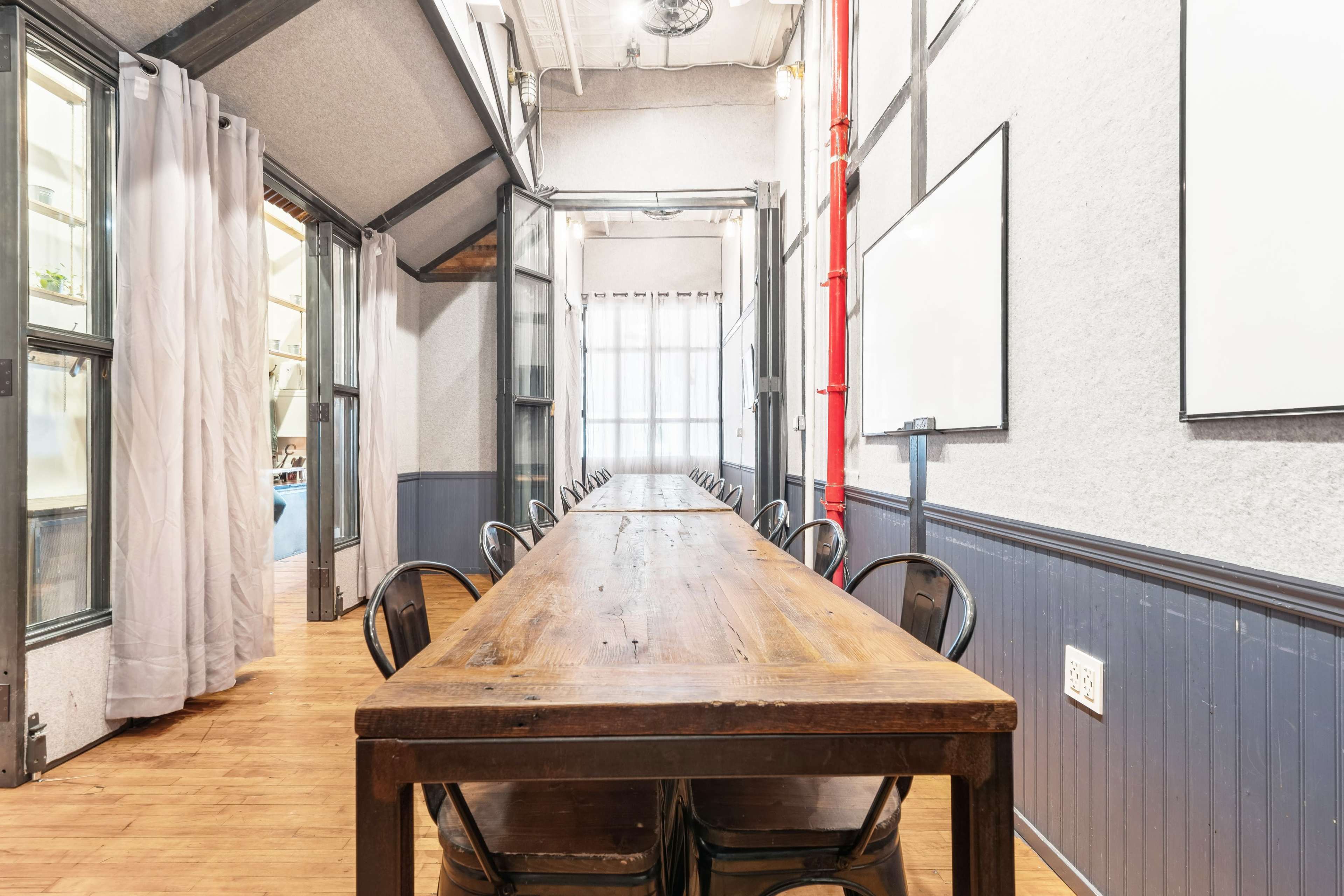 A long wooden table with black chairs is set in a well-lit room featuring gray walls and large windows.