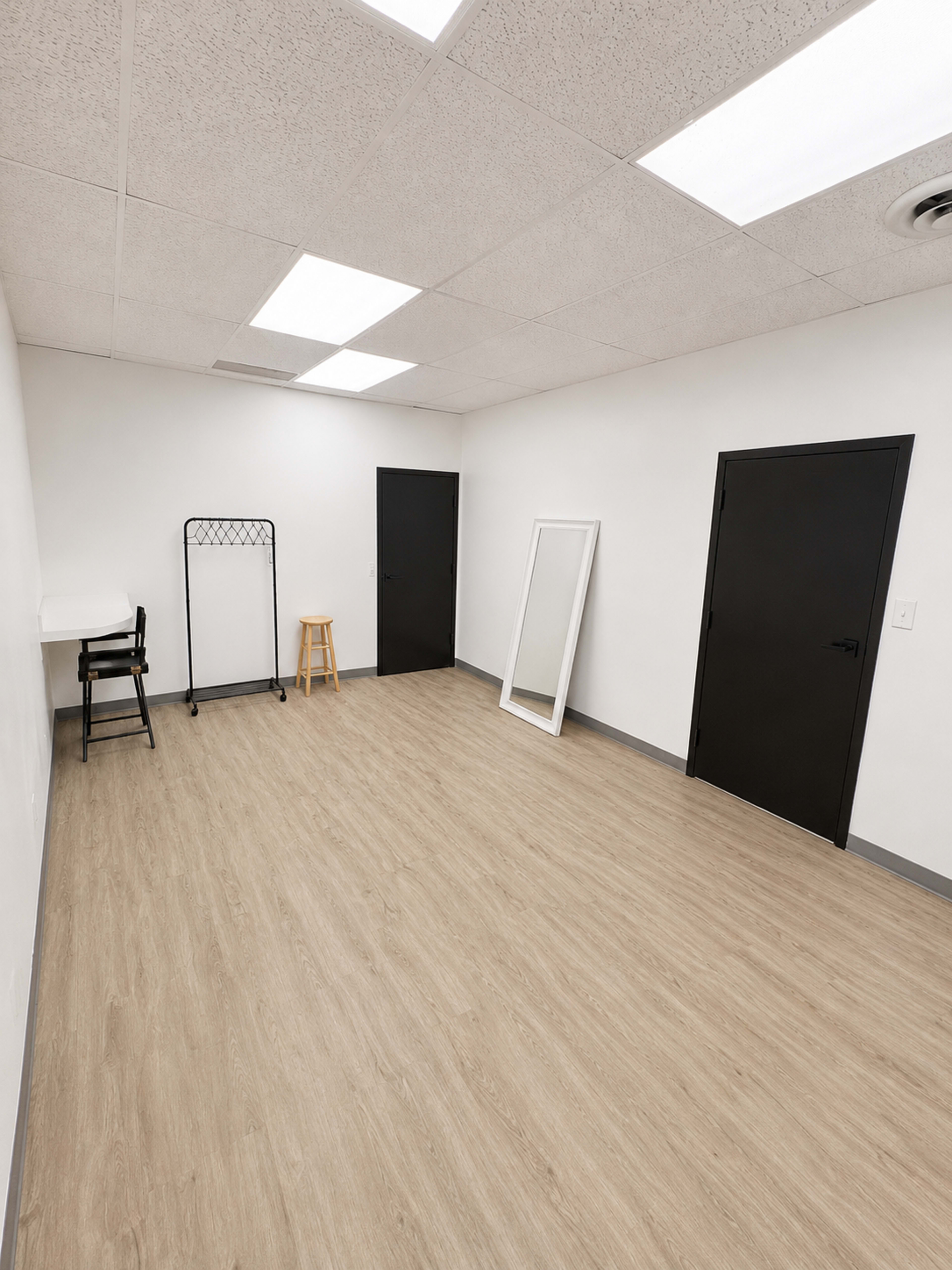 The image shows a sparsely furnished room with light wood flooring, a tall mirror, a clothing rack, and two doors, one of which is partially open.