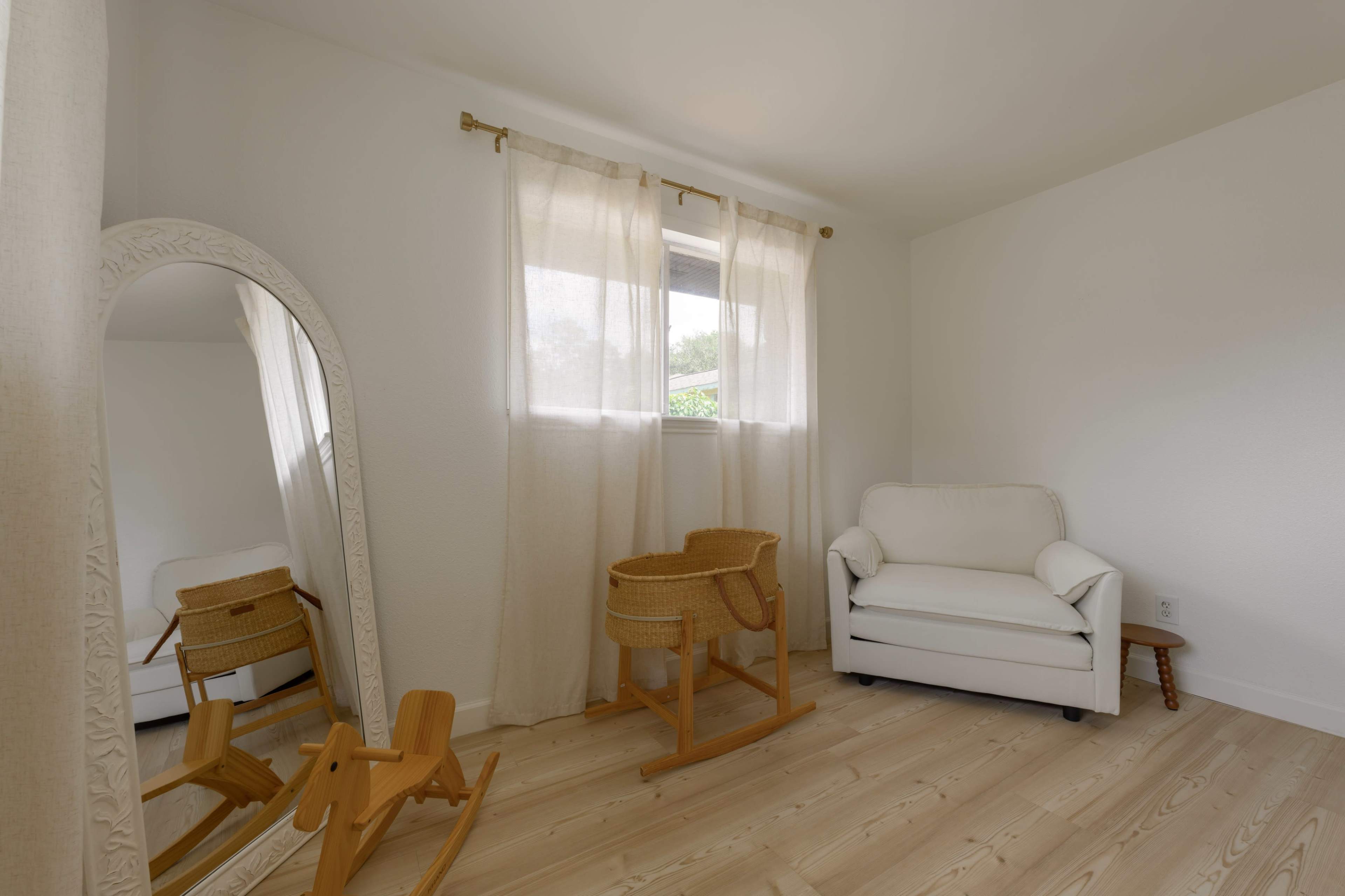 The image shows a bright, minimalistic room with a rocking chair, a white armchair, a large mirror, and sheer curtains covering a window.