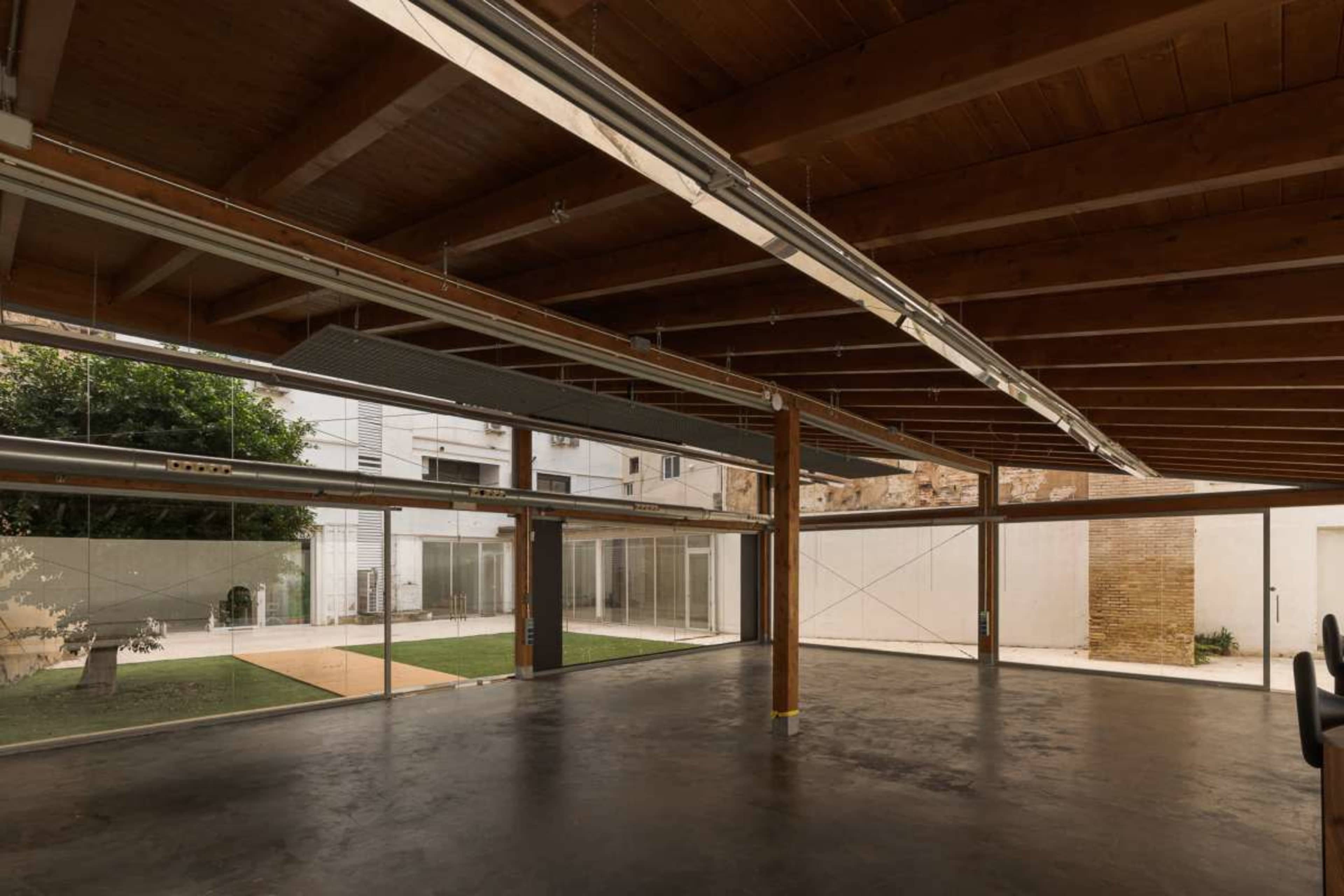 The image shows an open interior space with wooden beams, large glass windows, and a view of an outdoor area with grass and trees.