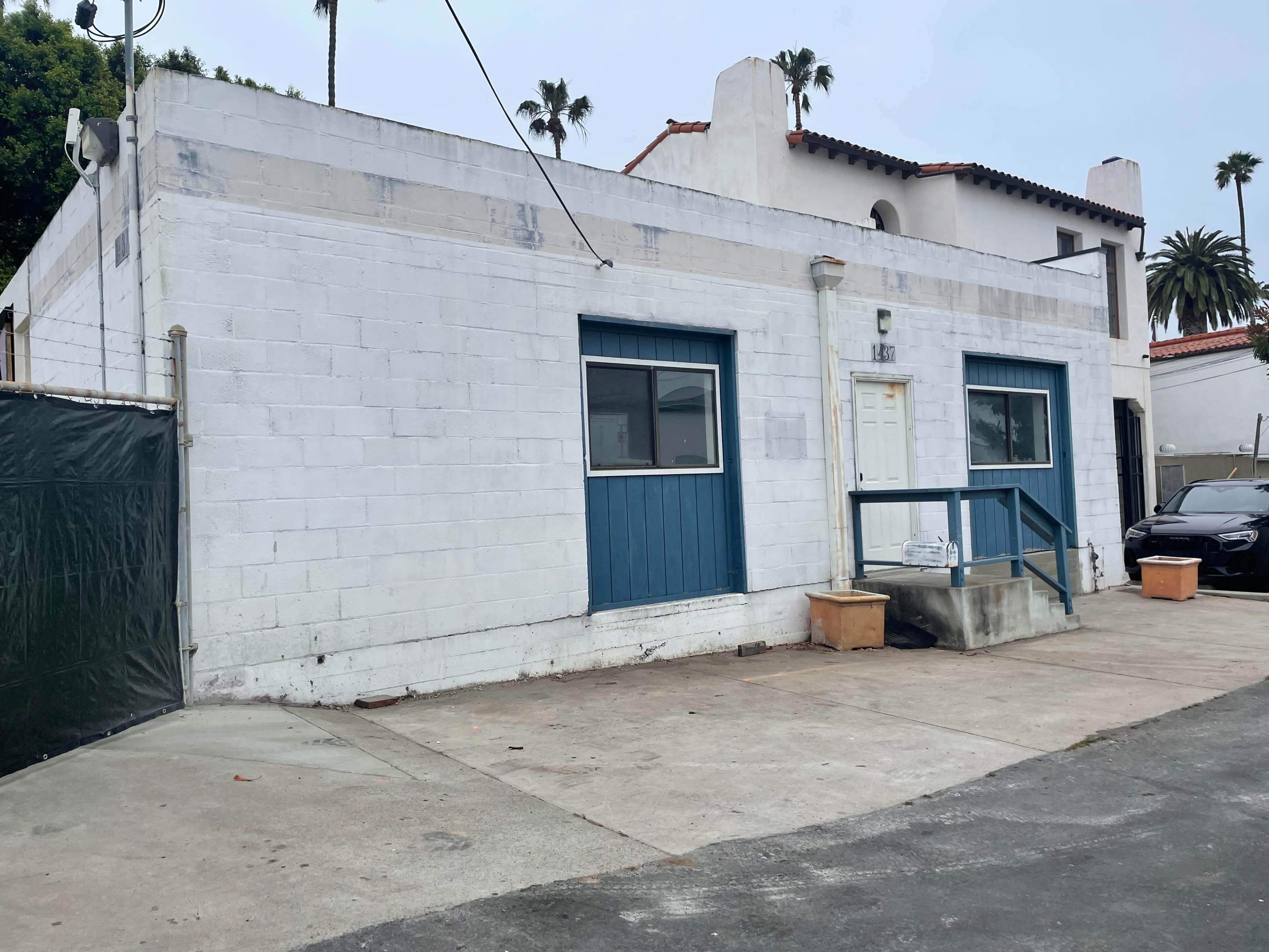 A white stucco building with a blue door and windows, set against a cloudy sky, surrounded by palm trees and pavement.