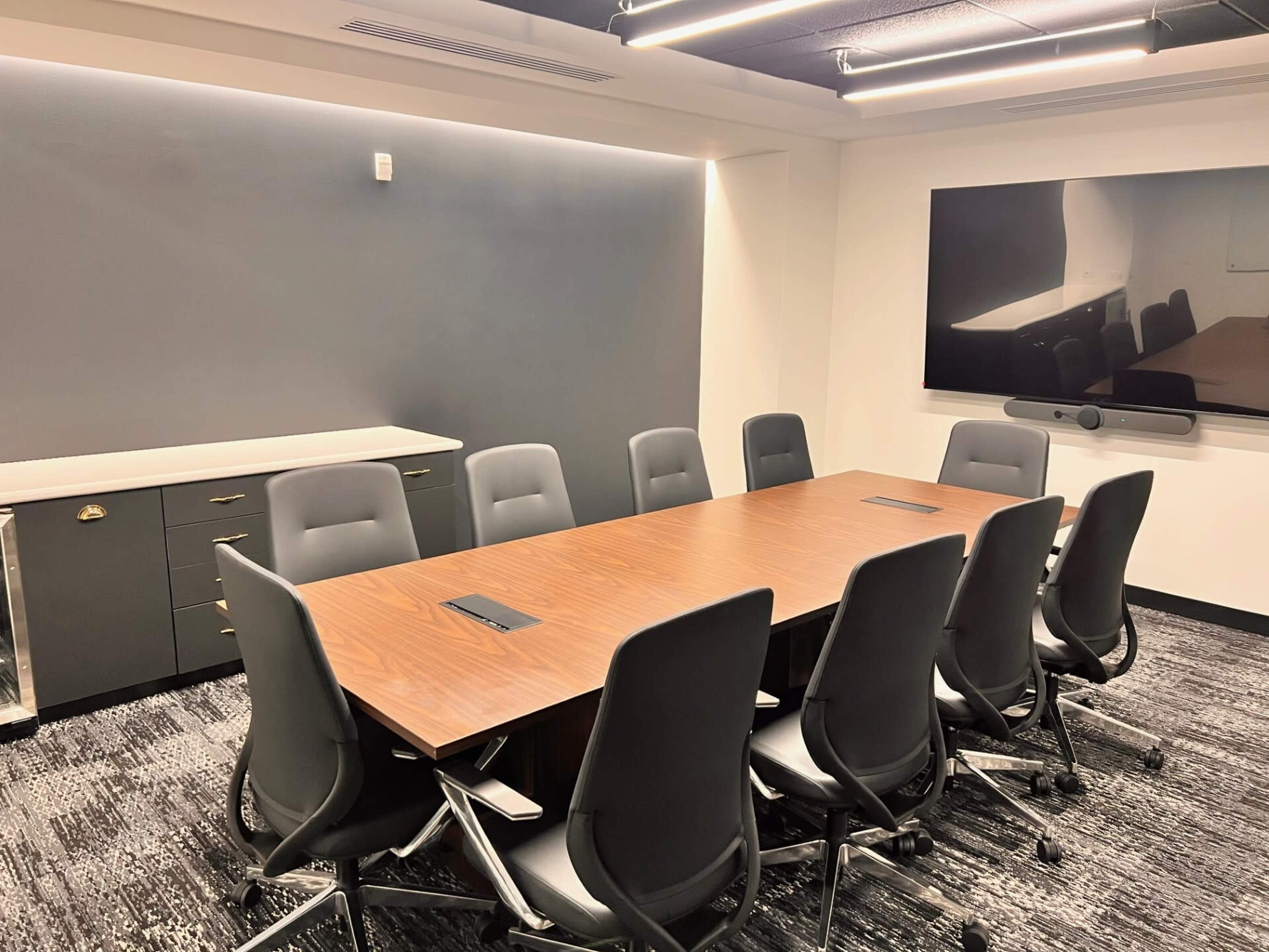 A modern conference room features a large wooden table surrounded by ten ergonomic office chairs, with a wall-mounted screen and minimal decor.