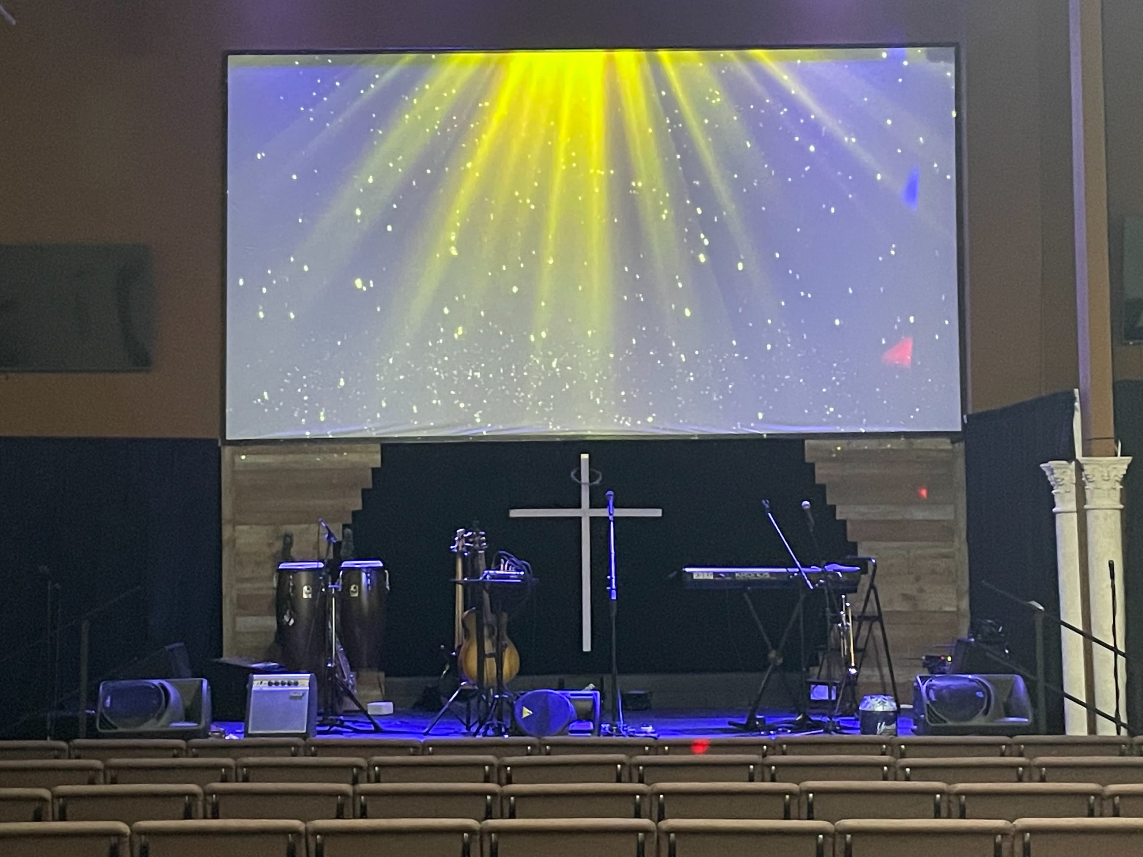 The image shows a stage in a church setting, featuring musical instruments like congas and a keyboard, with a large screen displaying beams of light in the background.