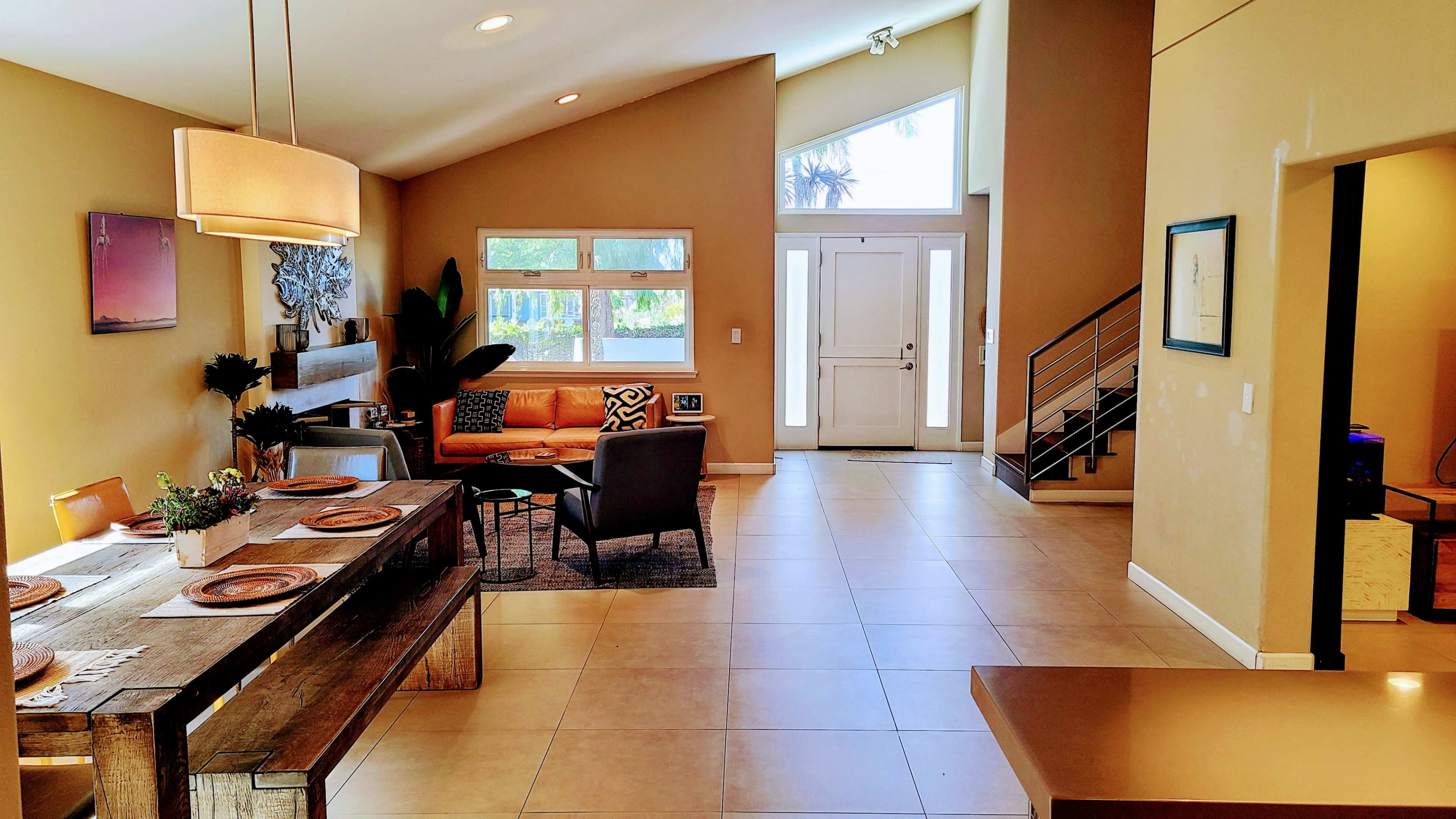 A modern, open-concept living space featuring a dining area with a wooden table, a cozy sitting area with a couch, and an entrance with large windows and a staircase.