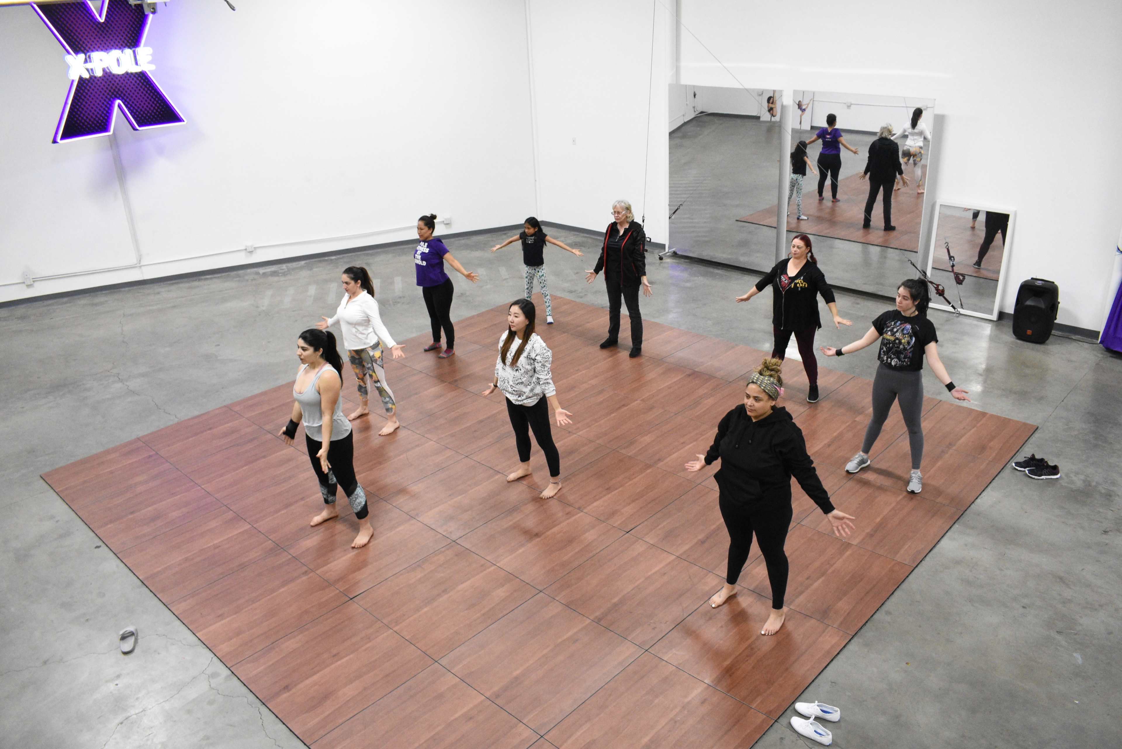 A group of women are participating in a dance class on a wooden floor in a well-lit studio with mirrors.