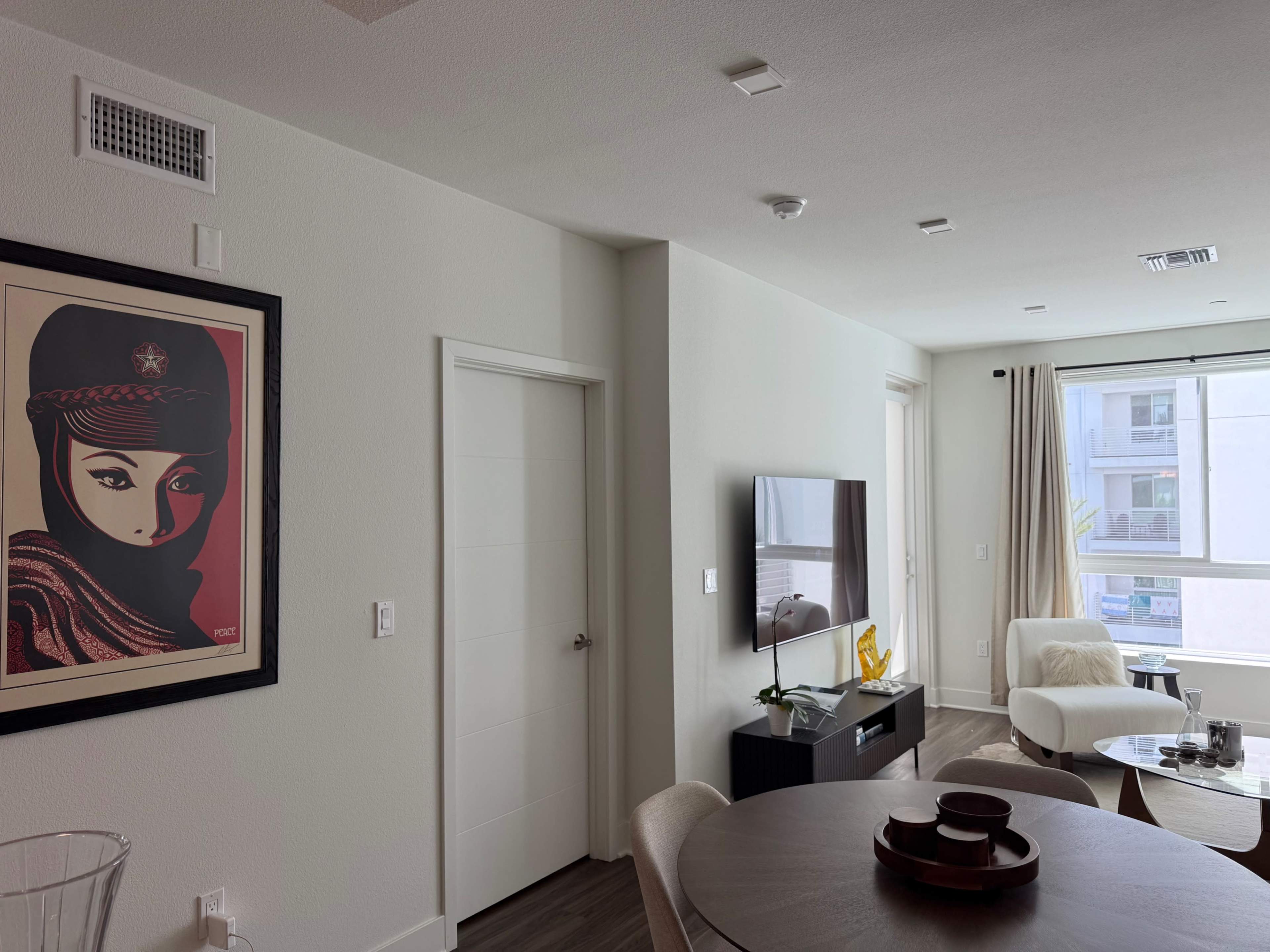 The image shows a modern living space featuring a wall-mounted television, a round dining table, and a wall decoration of a woman’s portrait.