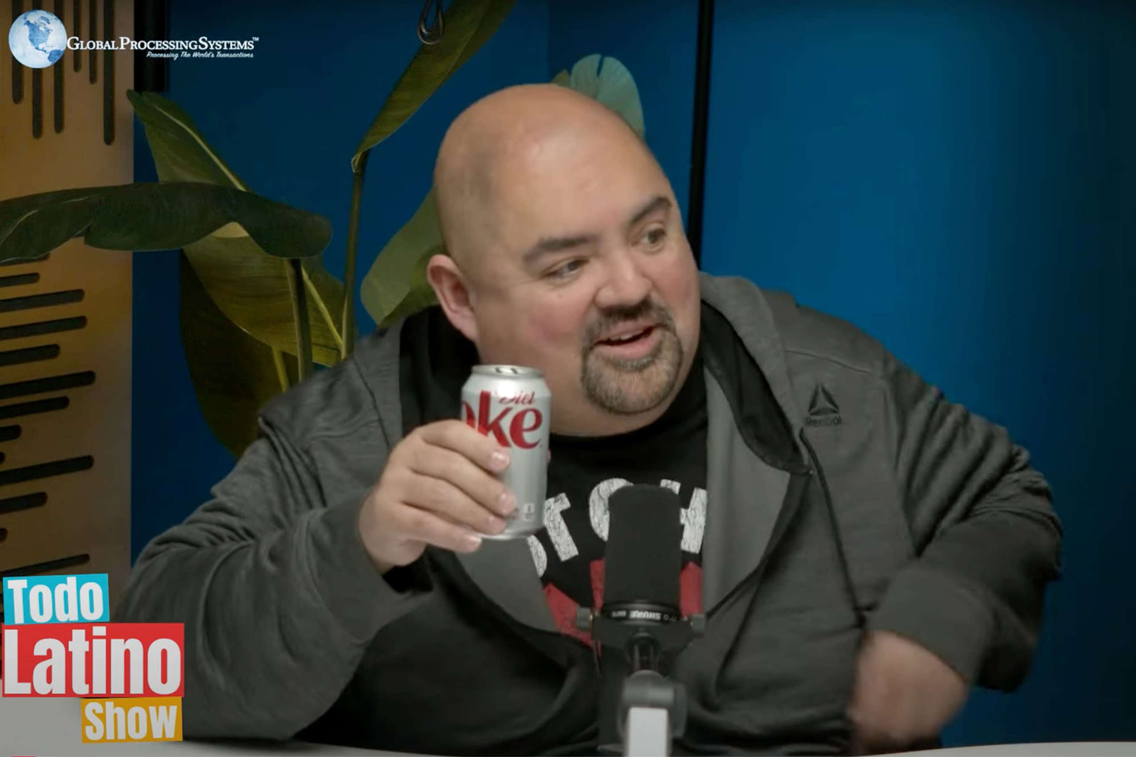 A man holding a can of Diet Coke sits at a table with a microphone in front of him, surrounded by green plants and a colorful backdrop.