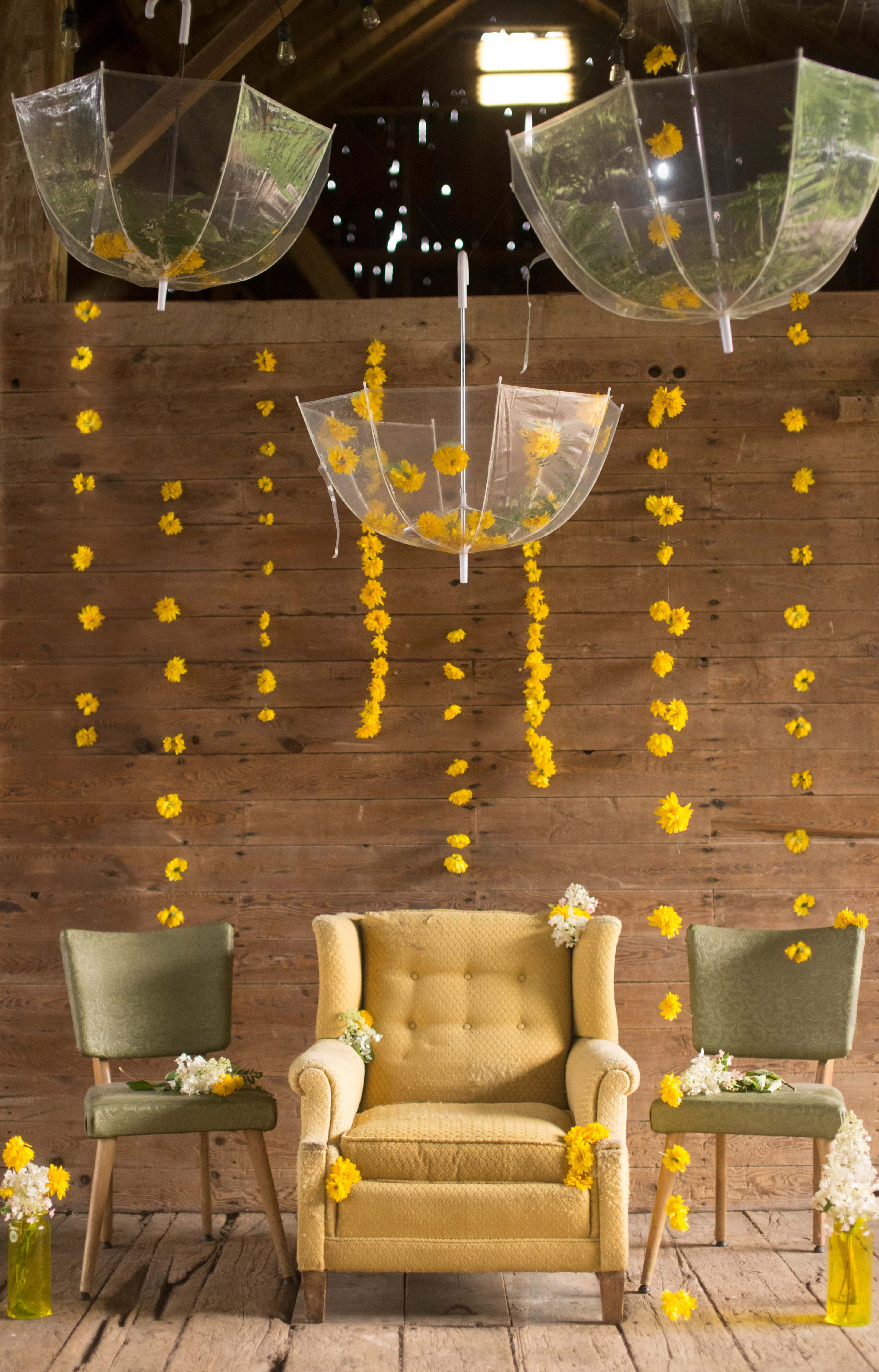 The scene features a rustic interior with a wooden wall adorned with hanging clear umbrellas and yellow flower decorations, alongside a yellow upholstered armchair and two green chairs.
