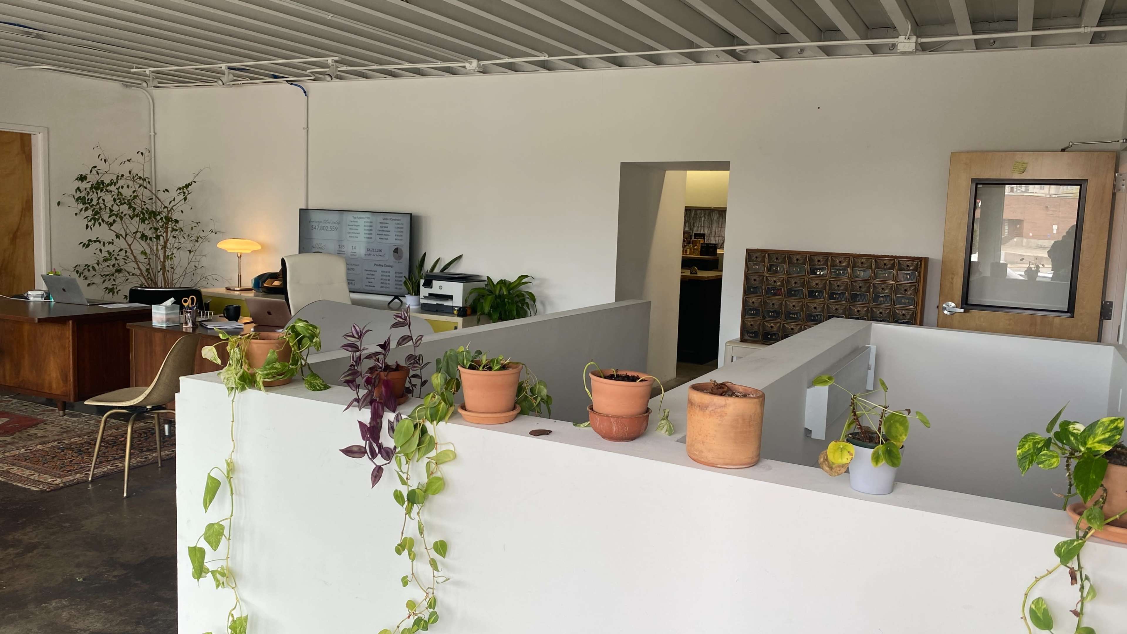 A modern office space with white walls, potted plants, desks, and a television on the wall.