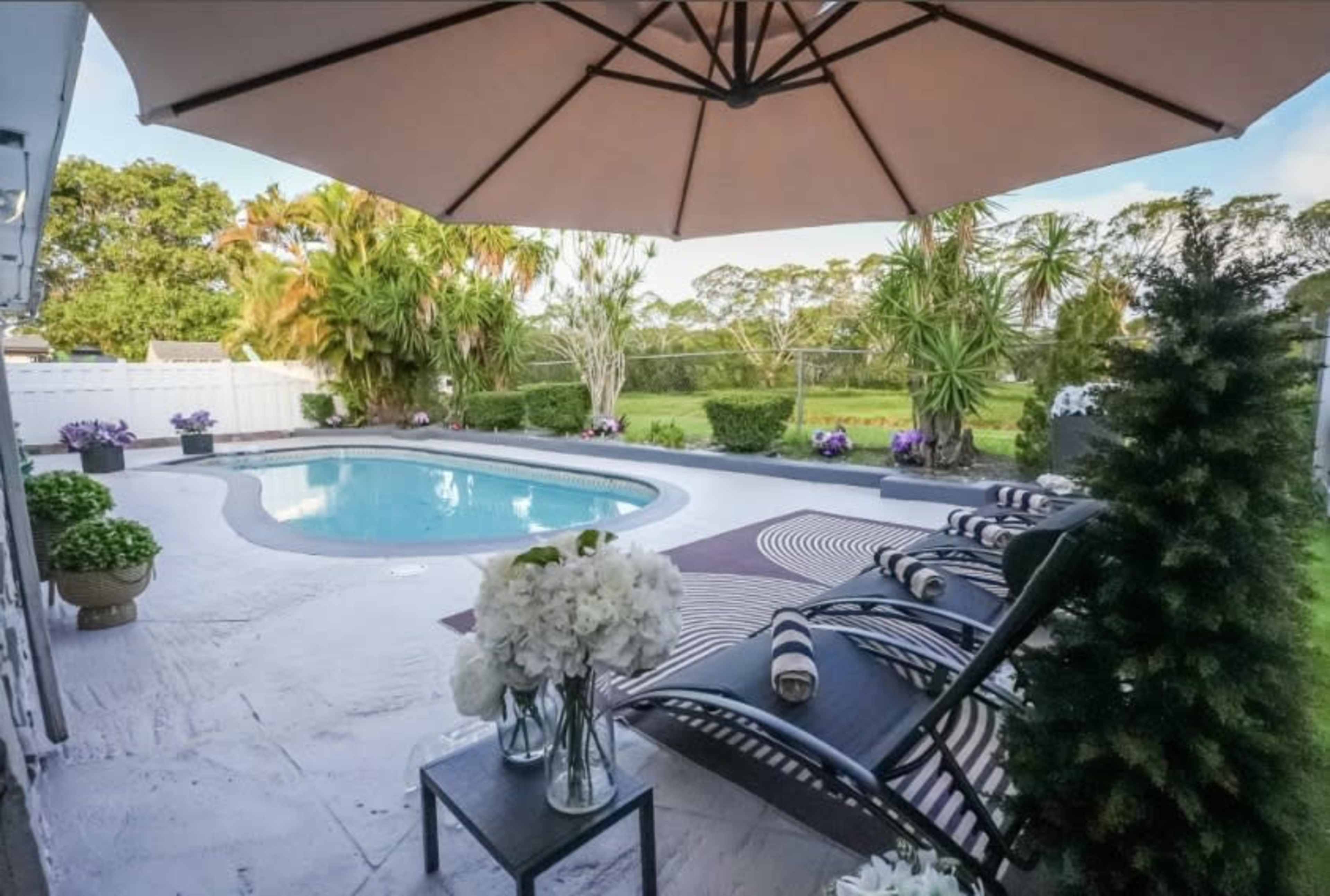 The image shows a backyard pool area with lounge chairs, a large umbrella, and surrounding tropical plants.