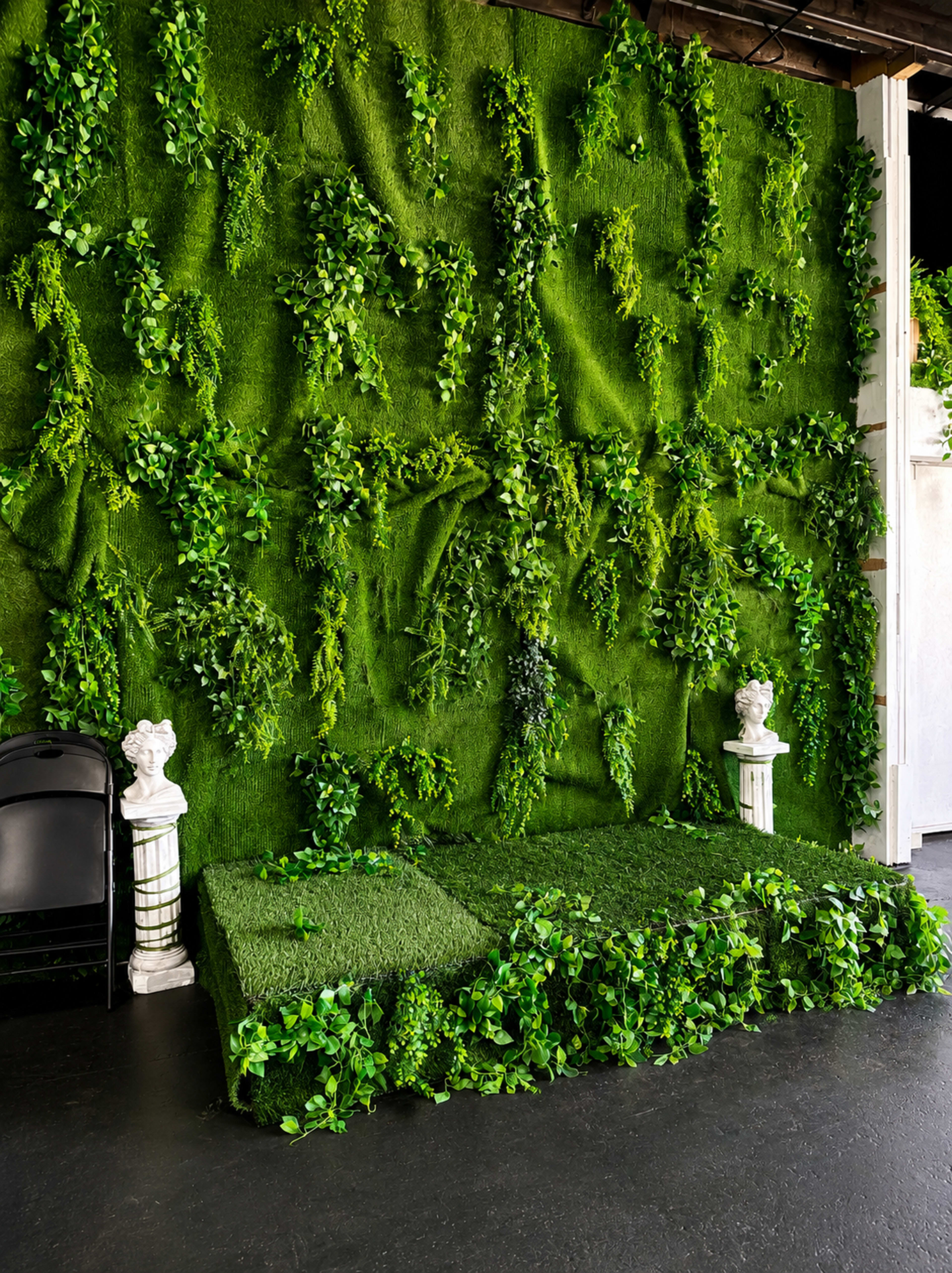 The image shows a green wall covered in artificial foliage with a grassy area in front, adorned with white statues on either side.
