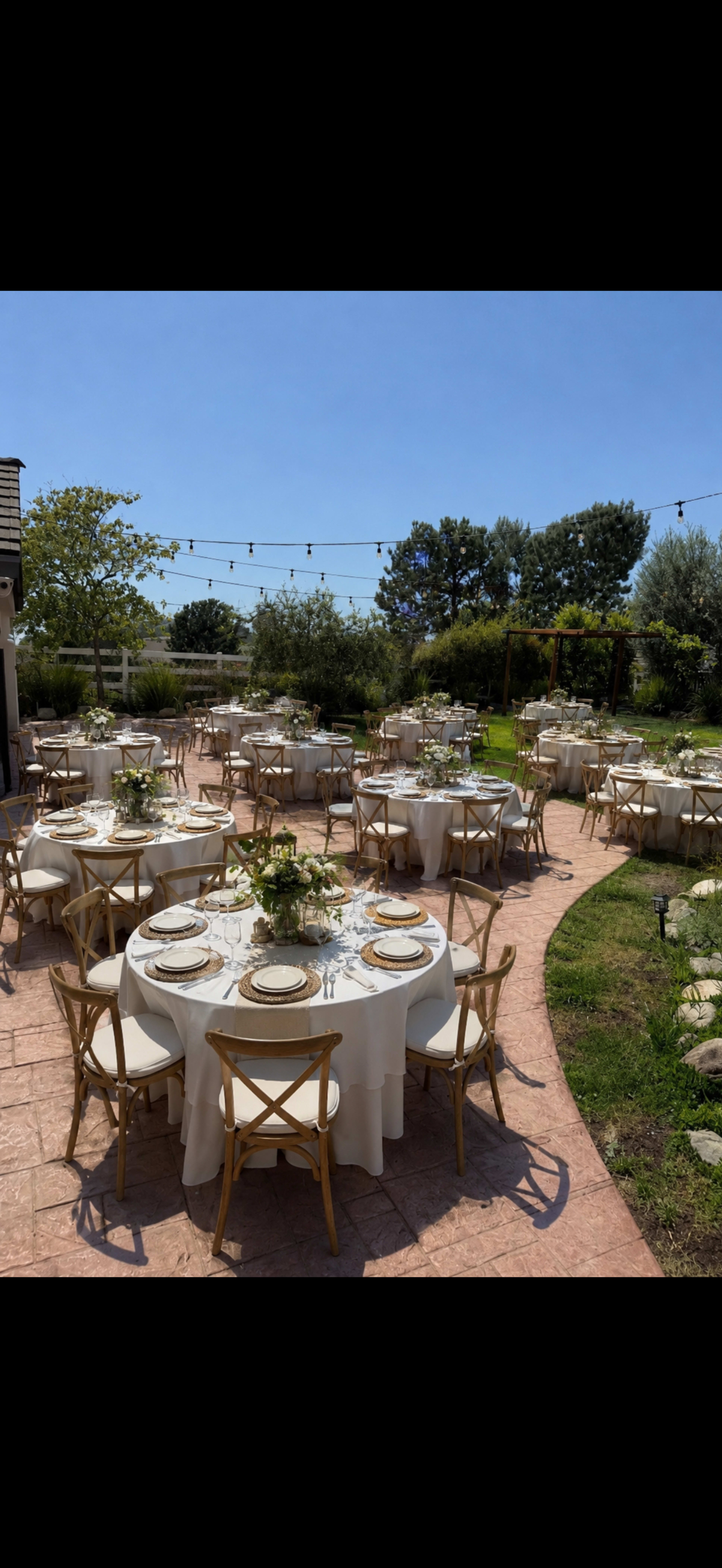 The image shows an outdoor dining area adorned with round tables set for a meal, surrounded by greenery and string lights overhead.