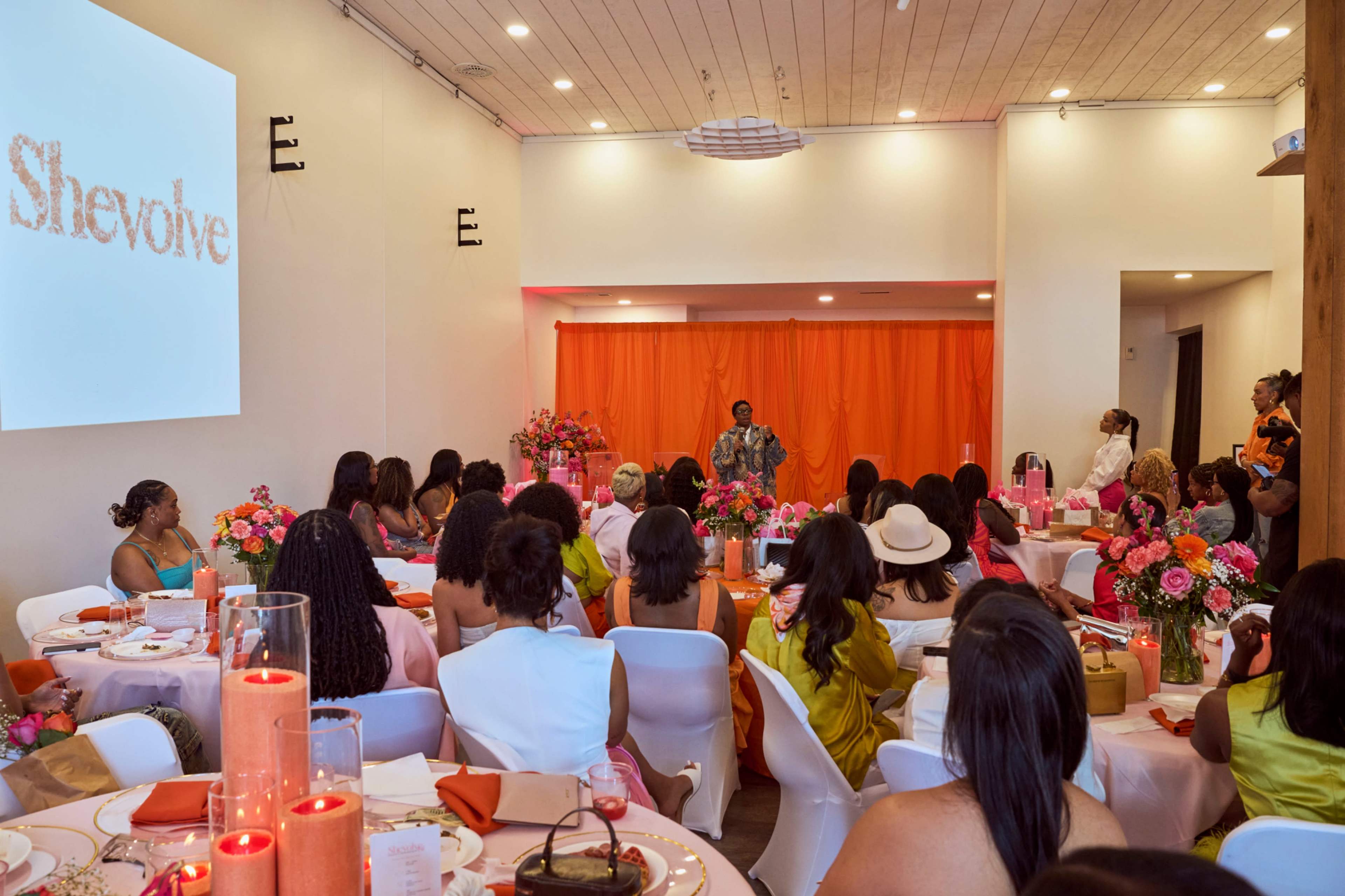 A speaker stands at the front of a room decorated with orange drapes and floral centerpieces, addressing an audience seated at tables arranged for an event.