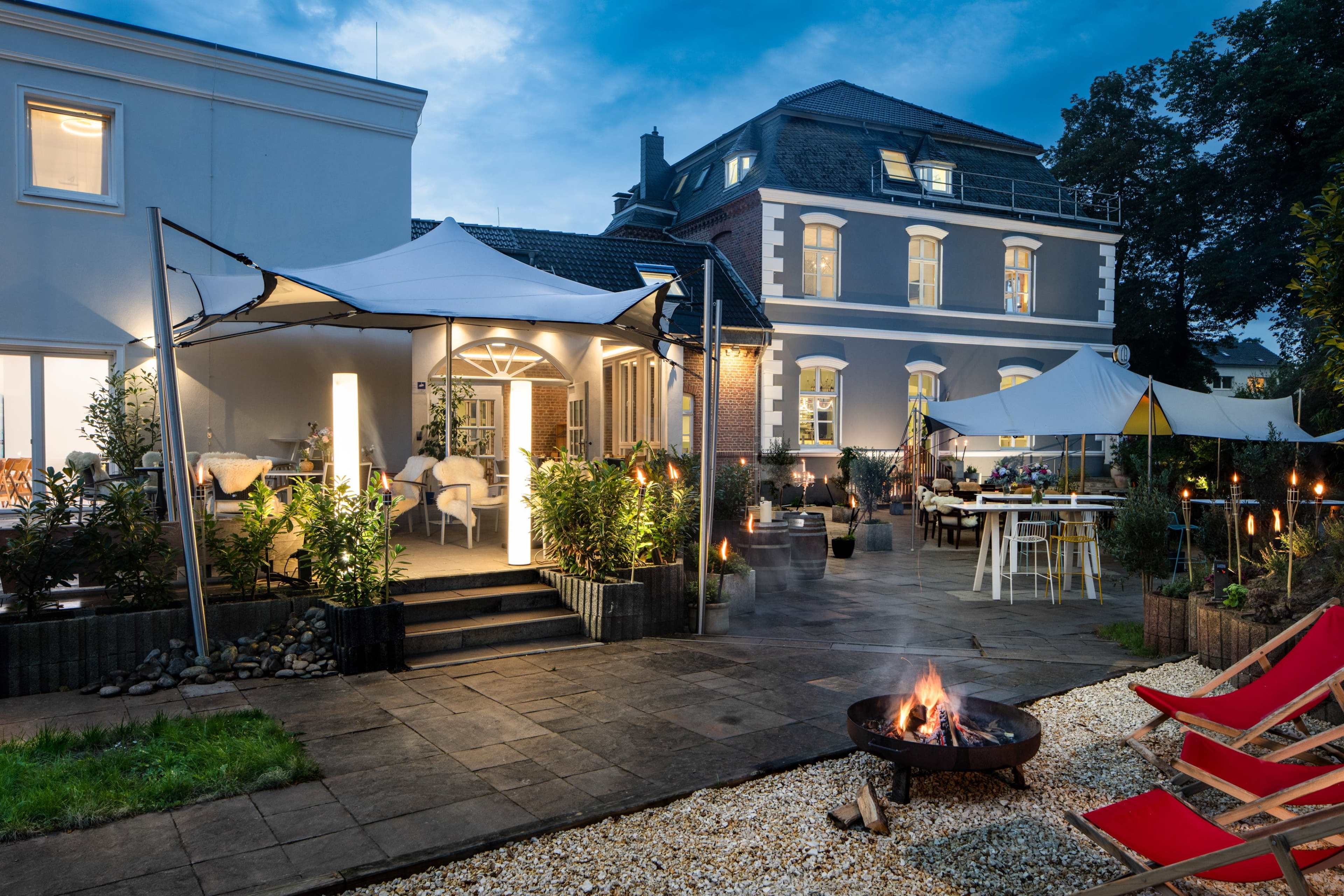 A well-lit outdoor seating area features tents, fire pit, and tables beside a large building at twilight.