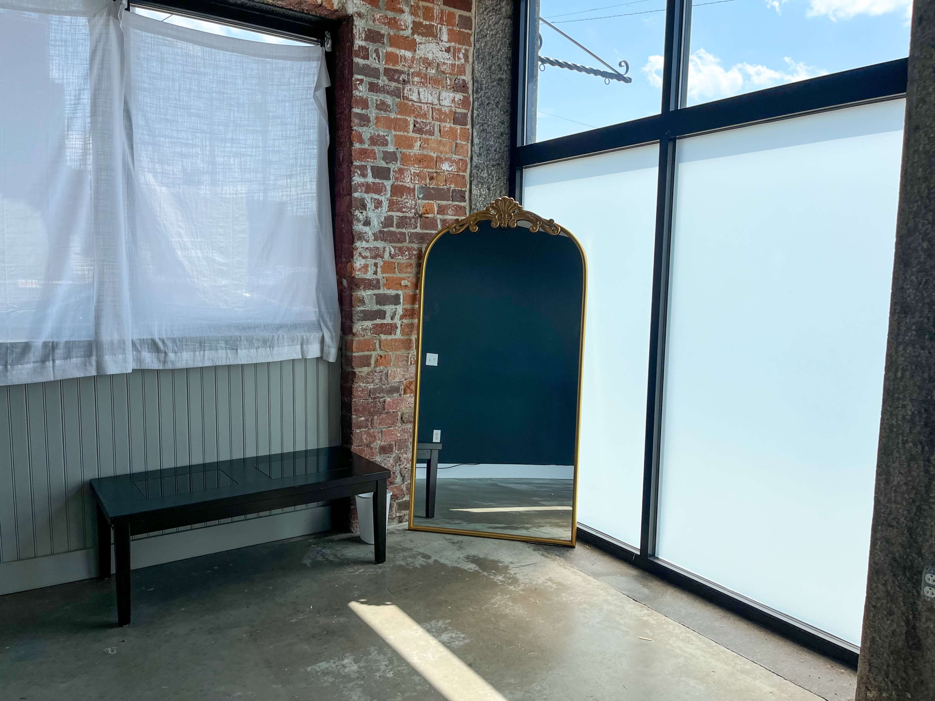 A large, ornate mirror stands against a wall beside a black bench in a room with brick and windowed walls.