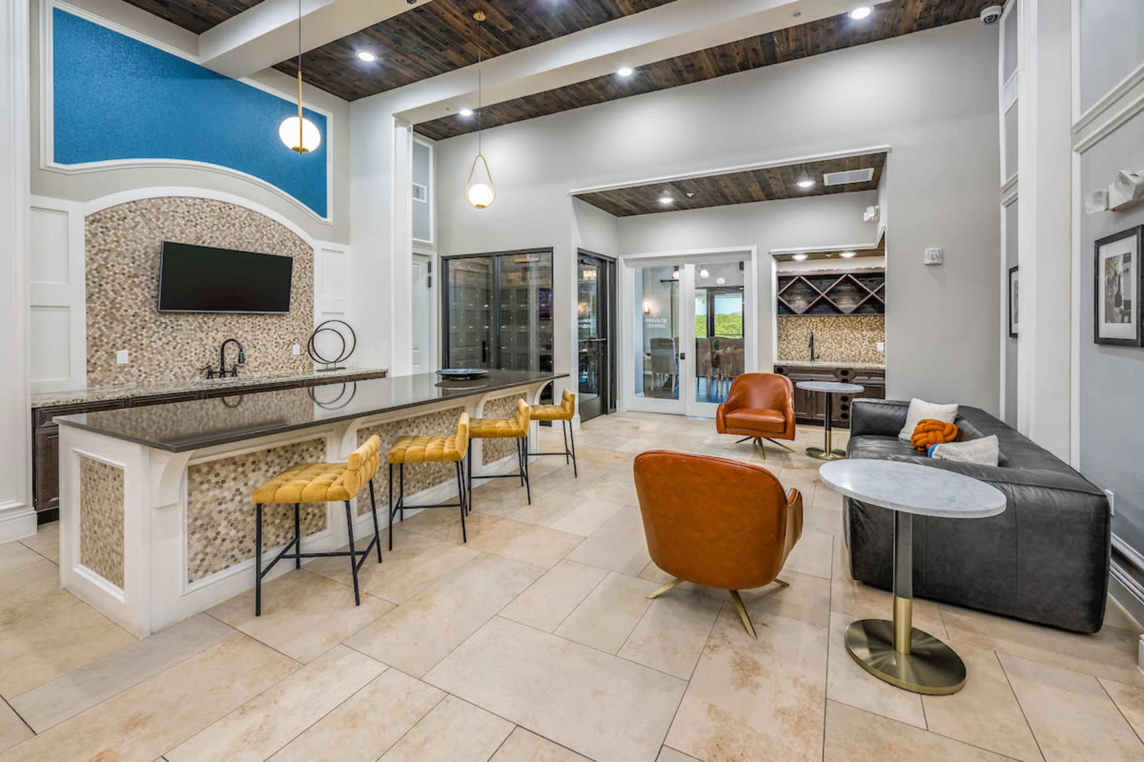 The image shows a modern lounge area featuring a large countertop with bar stools, a seating arrangement with leather chairs, and a glass-enclosed wine cellar in the background.