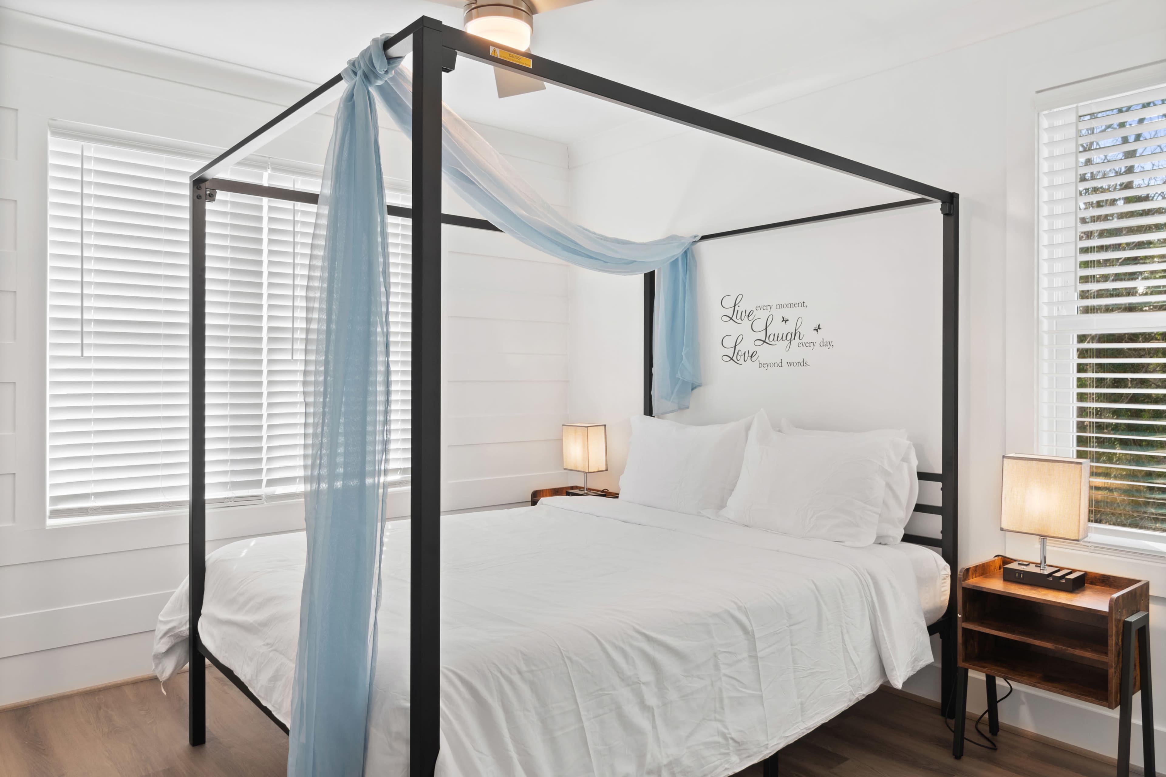 A four-poster bed with white linens is positioned against a wall in a bright room featuring wooden flooring and two bedside lamps.