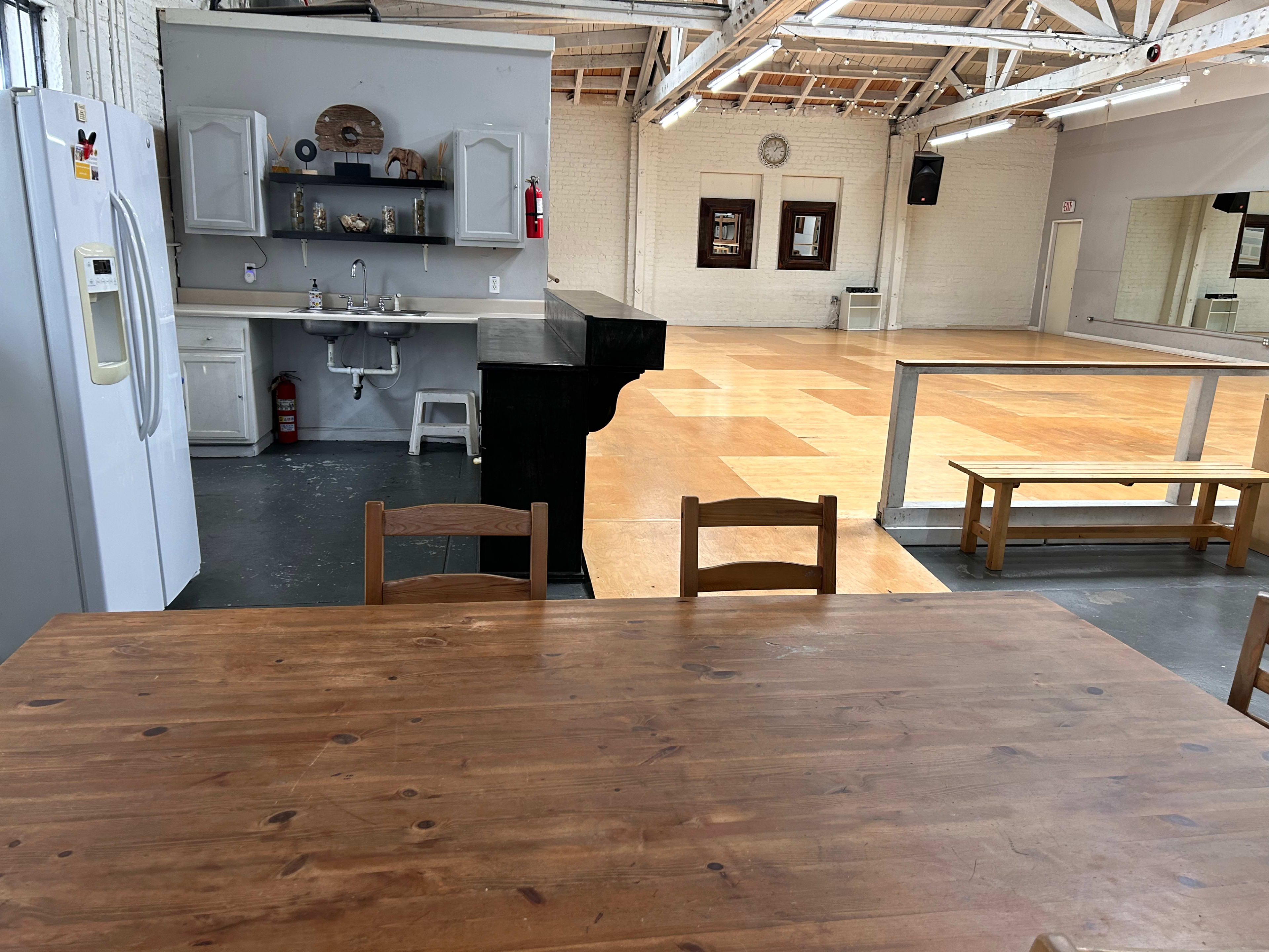 An empty wooden floor studio with a kitchen area featuring cabinets, a sink, and a refrigerator in the background.