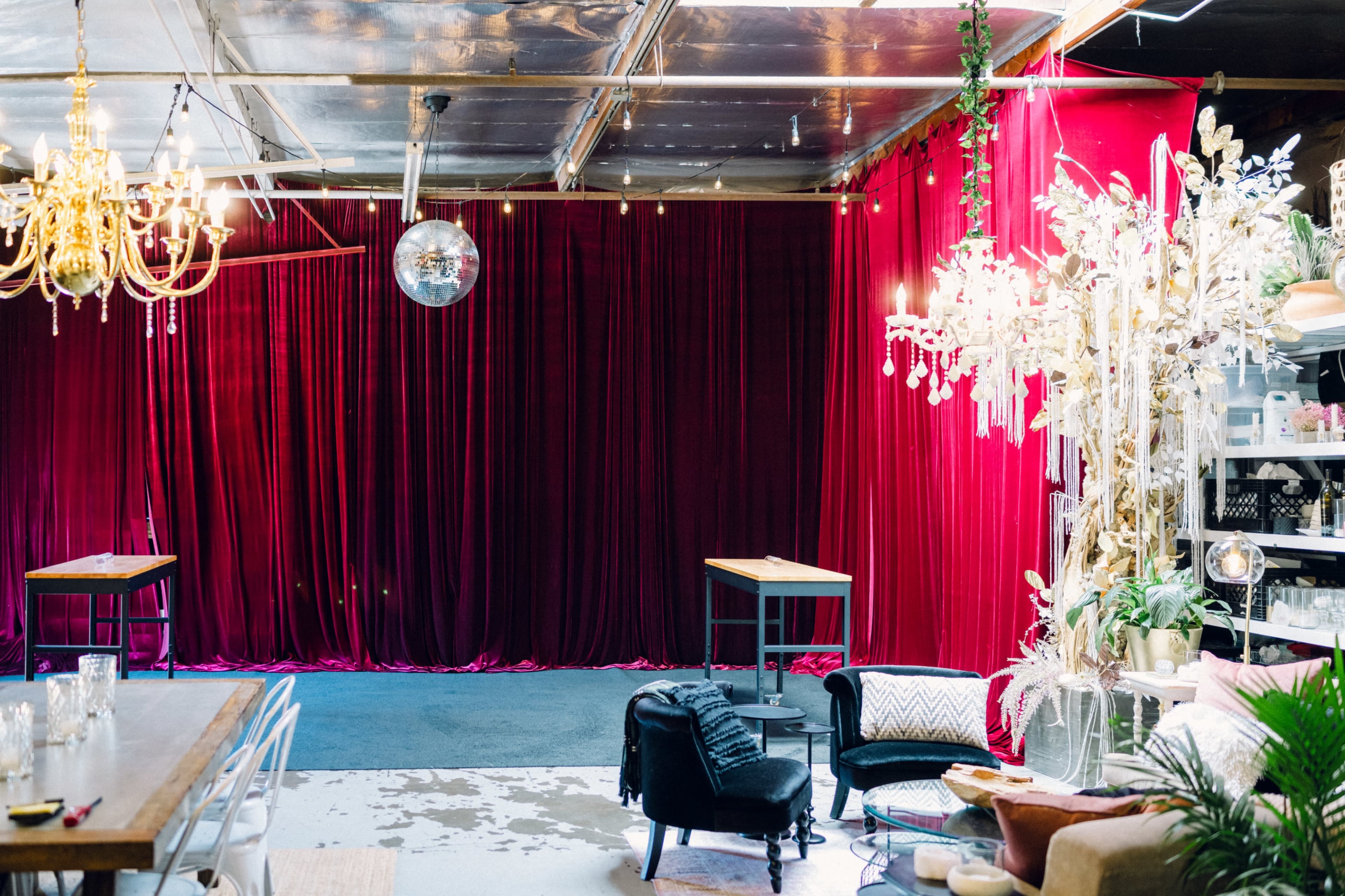 The image shows a spacious indoor venue with a deep red velvet backdrop, two wooden tables, and decorative chandeliers overhead.