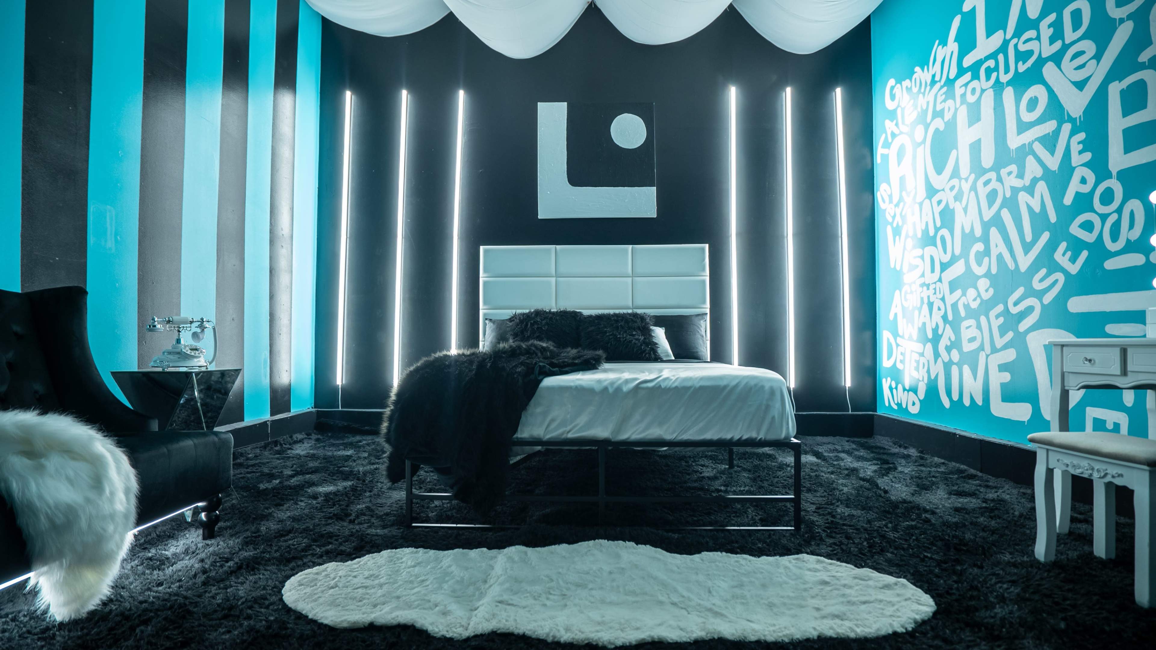 The image shows a modern bedroom with a bed covered in white linens, a black shag carpet, and walls painted in contrasting teal and black, featuring decorative lighting and text art.