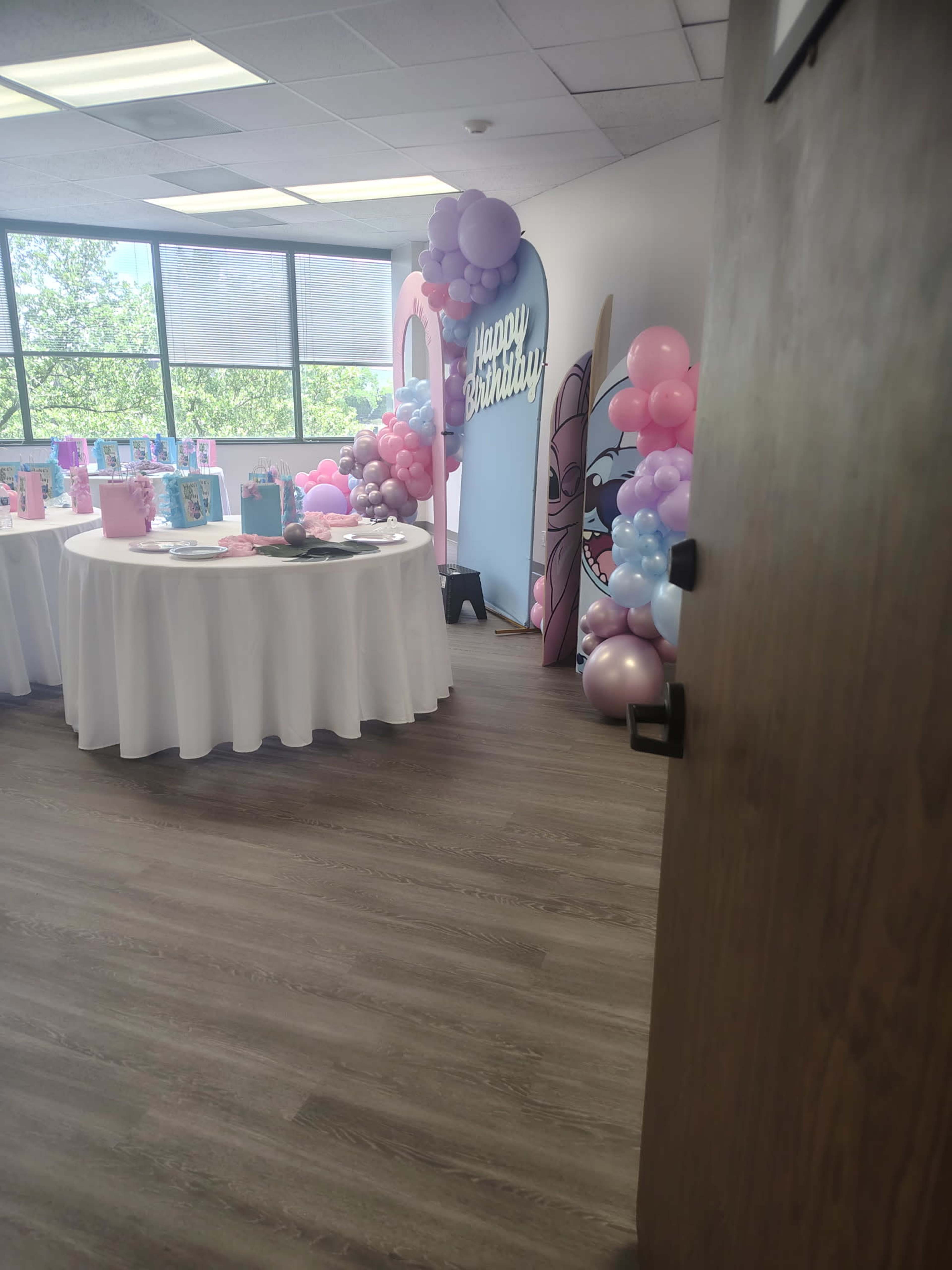 The image shows a decorated birthday celebration setup inside a room, featuring tables covered with white tablecloths and colorful balloons.