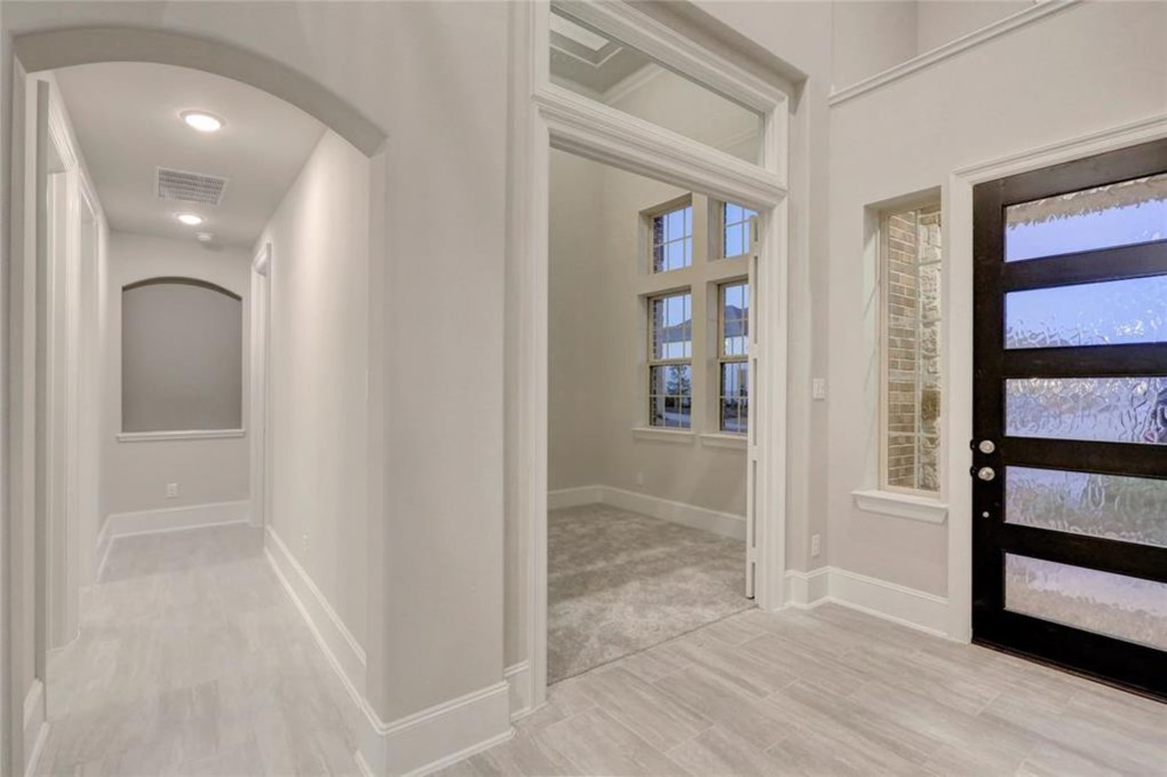 An interior entryway with a hallway leading to a room, featuring large windows and a modern front door with frosted glass panels.