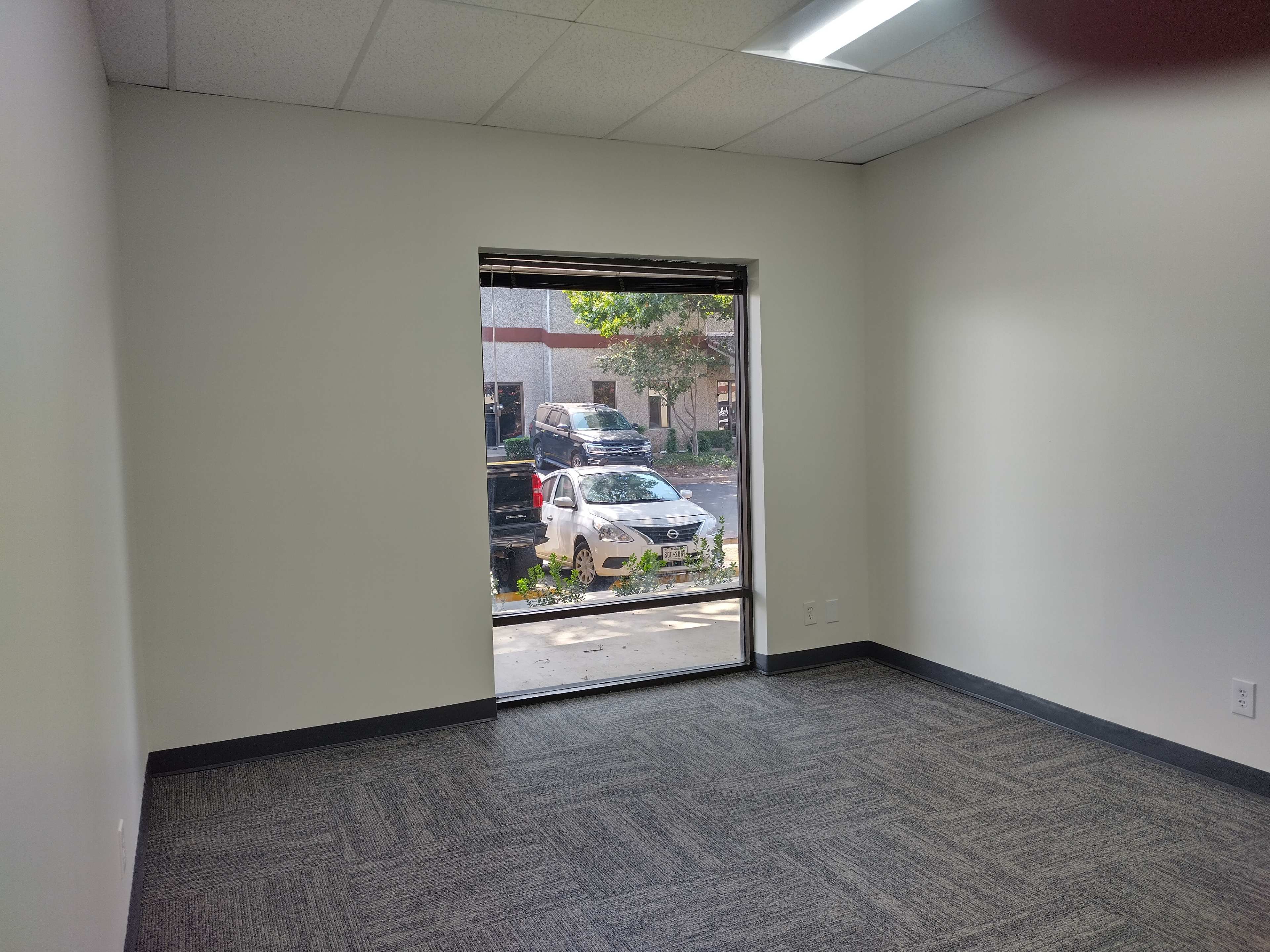 The image shows an empty room with gray carpet and a large window looking out onto a parking area with parked cars.