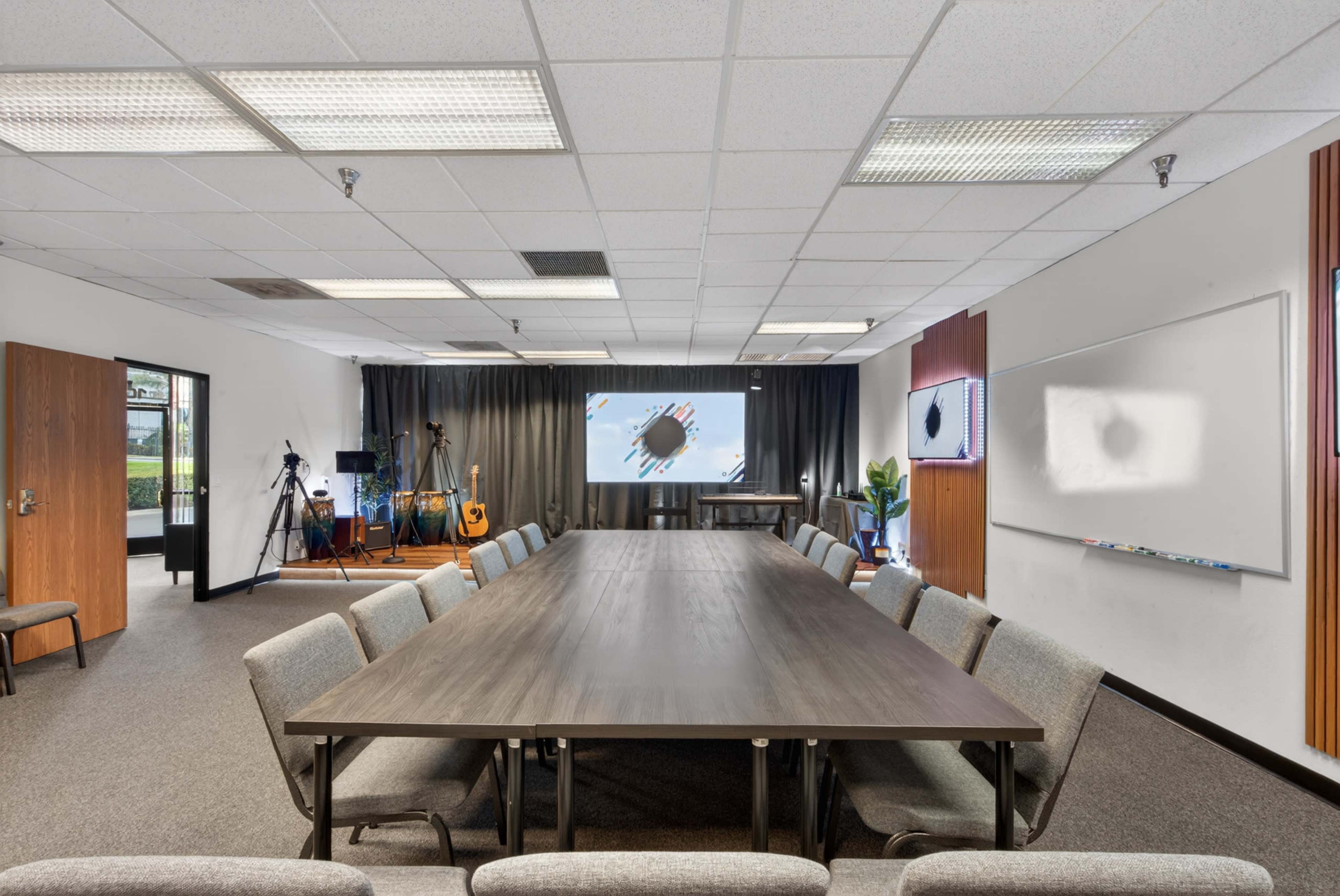 The image shows a large conference room with a long table surrounded by chairs, a projector screen on one wall, and equipment for video recording in the background.