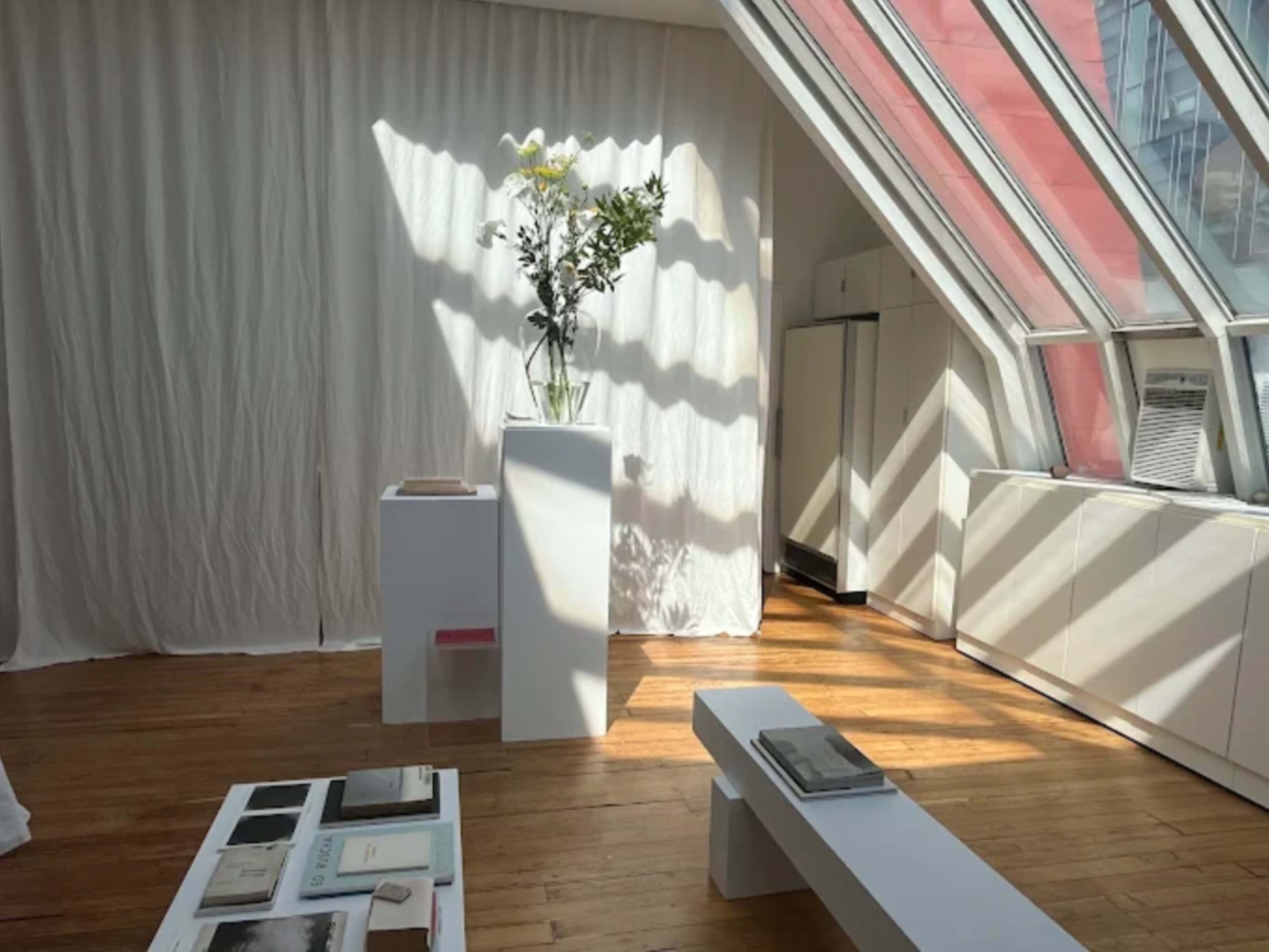The image shows a bright, airy interior space with hardwood floors, large windows, and a white pedestal displaying a vase of flowers.