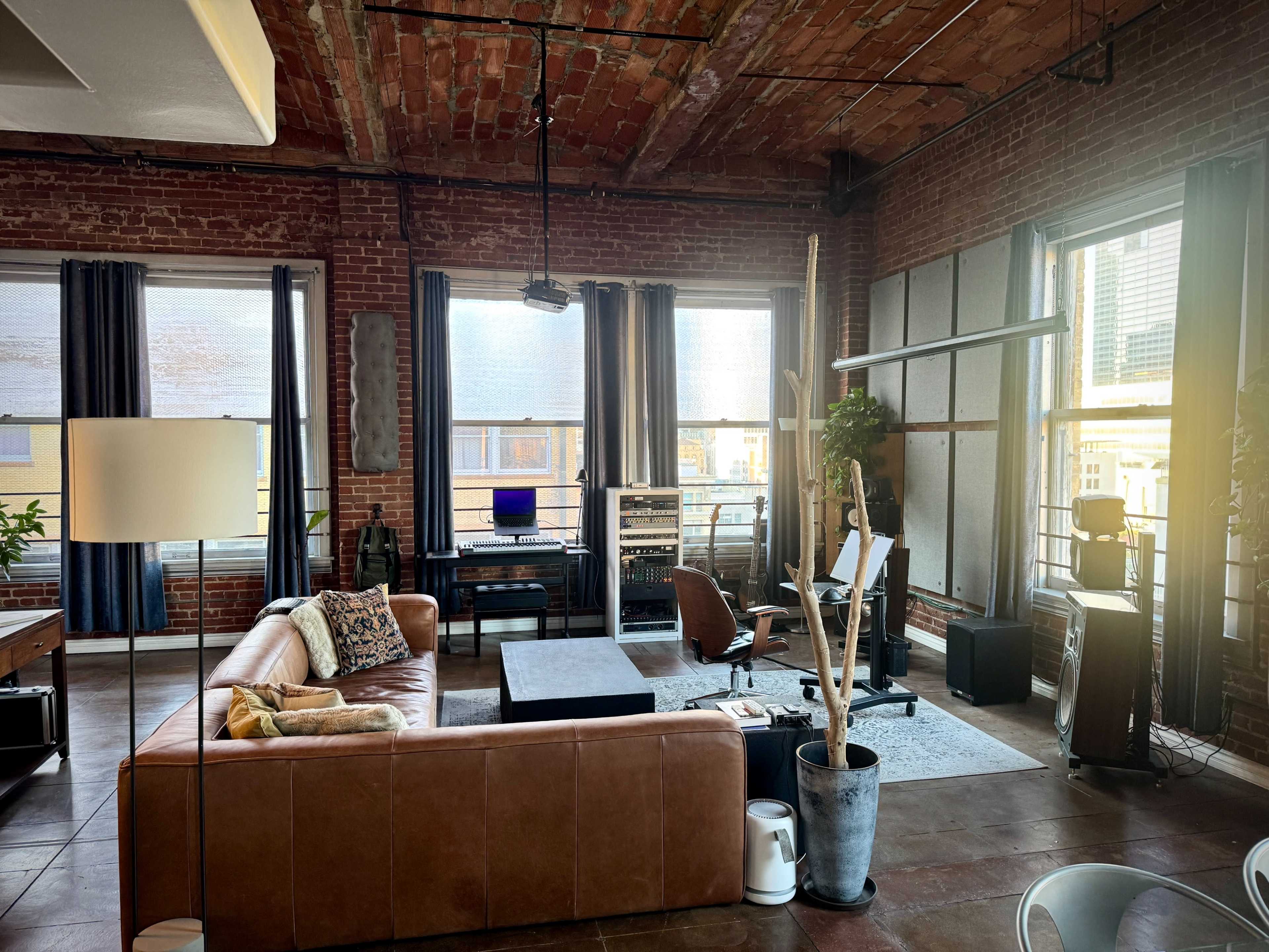 The image shows a spacious industrial-style living room featuring exposed brick walls, large windows, a brown leather sectional sofa, a coffee table, and a desk with a computer setup.