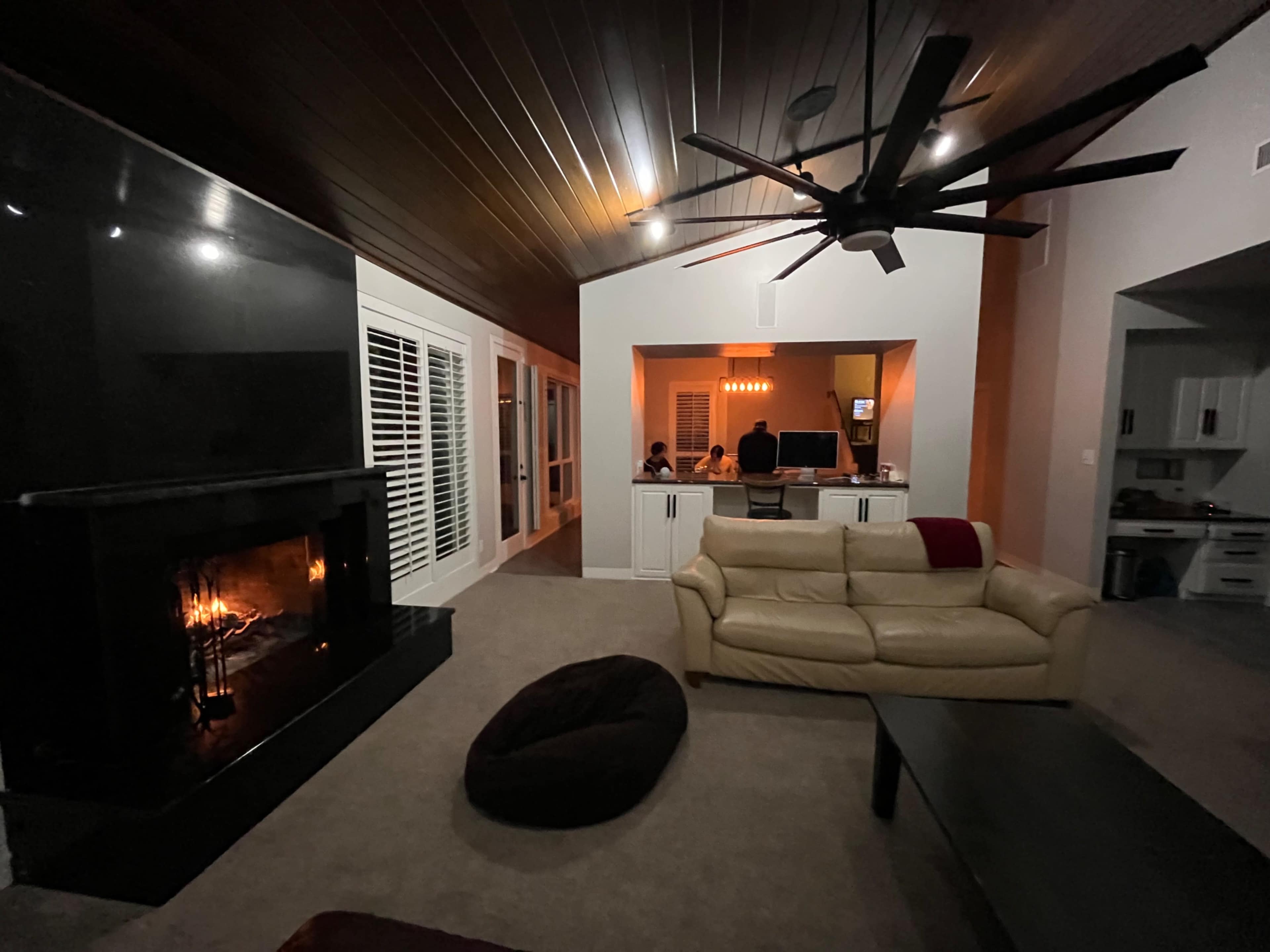 The image shows a cozy living room with a fireplace, a beige couch, and a dark coffee table, while a dining area with people is visible in the background.