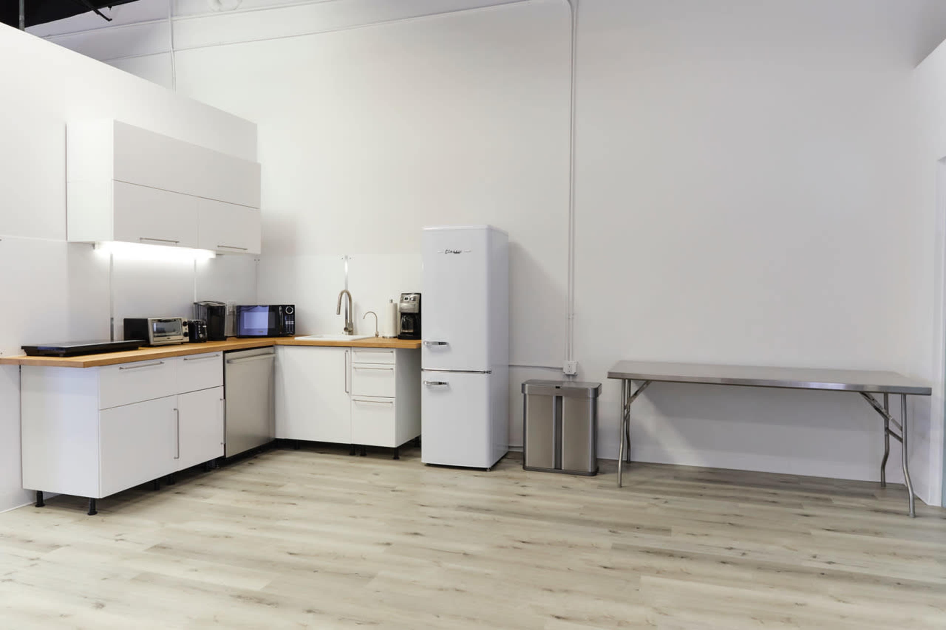 A modern kitchen area featuring a refrigerator, sink, and countertop appliances against a white wall, with a stainless steel table nearby.