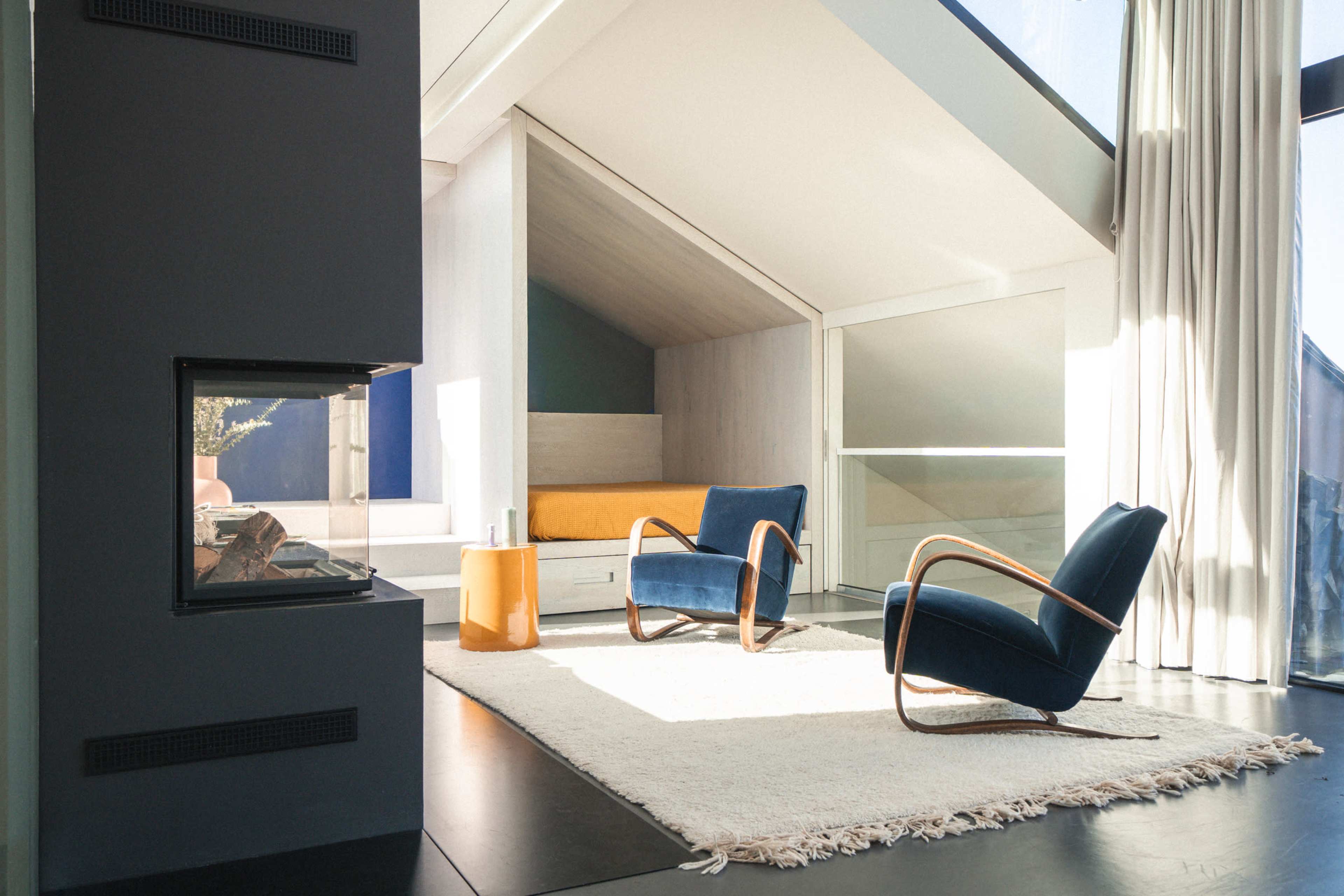 The image features a modern interior design with two blue chairs, a small orange table, a fireplace, and a bed in a cozy nook beneath a slanted ceiling.