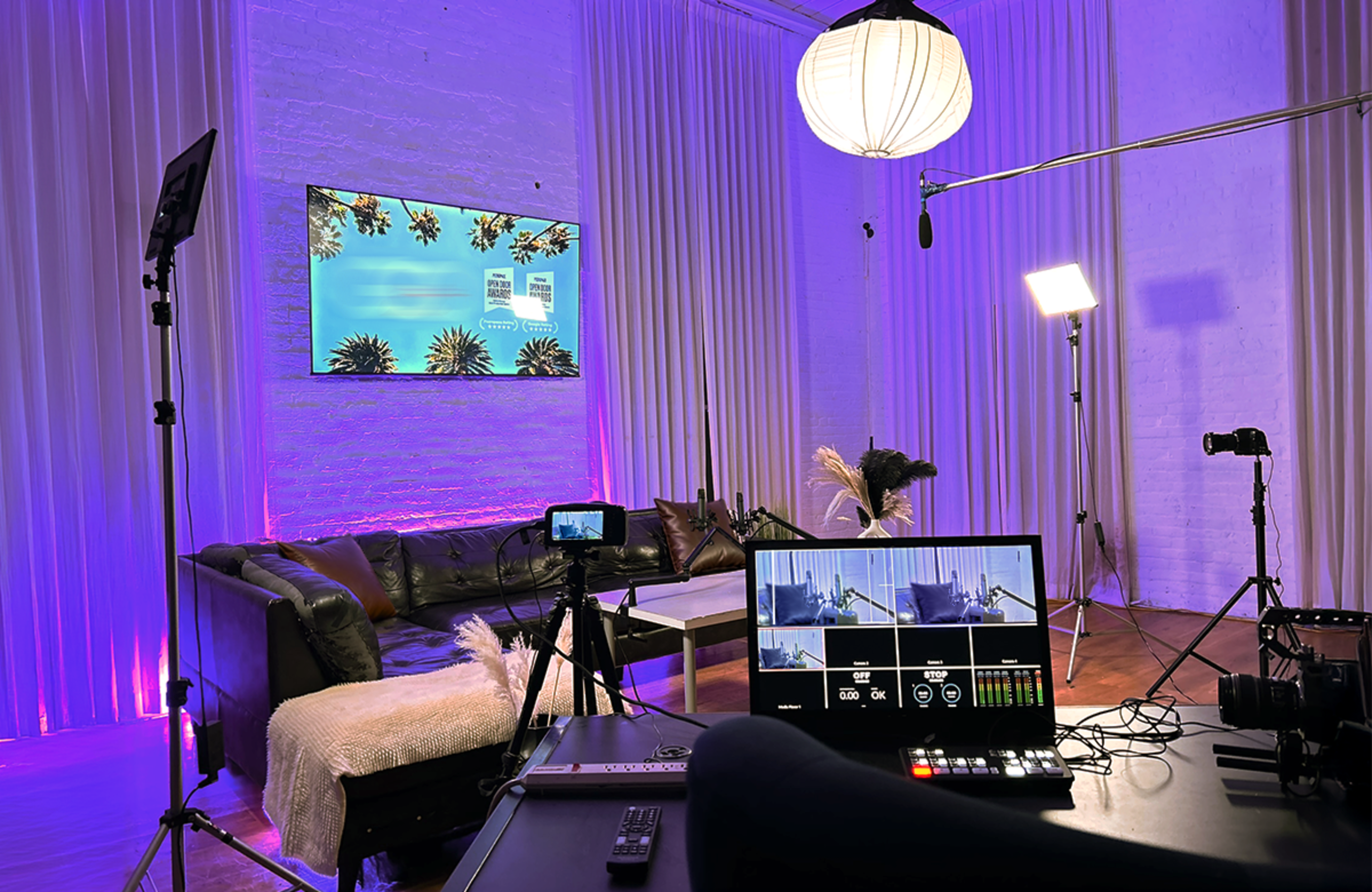 The image shows a modern video production setup with a camera, a monitor displaying visuals, and a well-lit lounge area featuring a black couch and decorative elements.