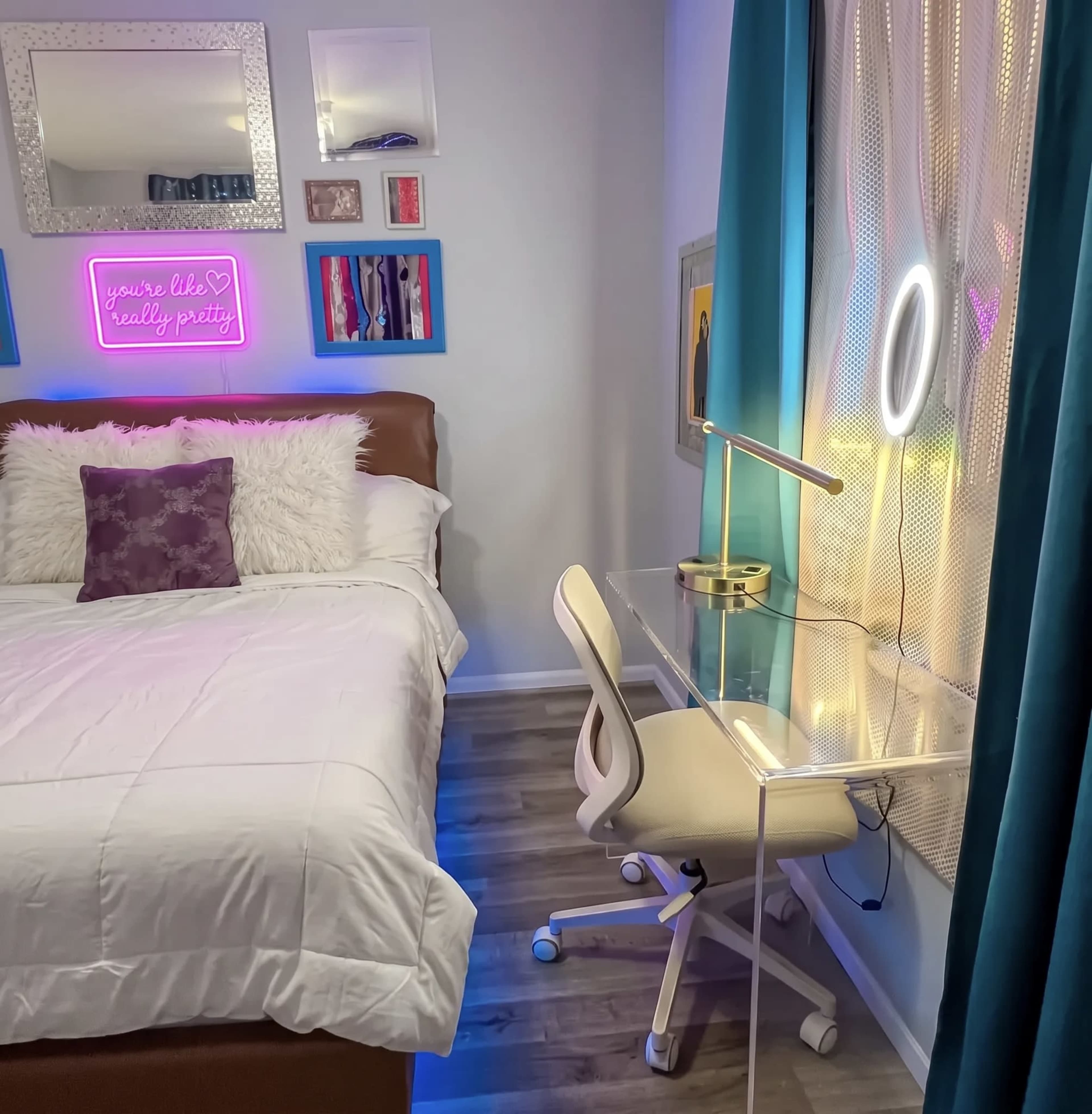 The image shows a bedroom featuring a bed with white bedding, a clear desk with a lamp, and a window with sheer curtains and decorative wall frames.
