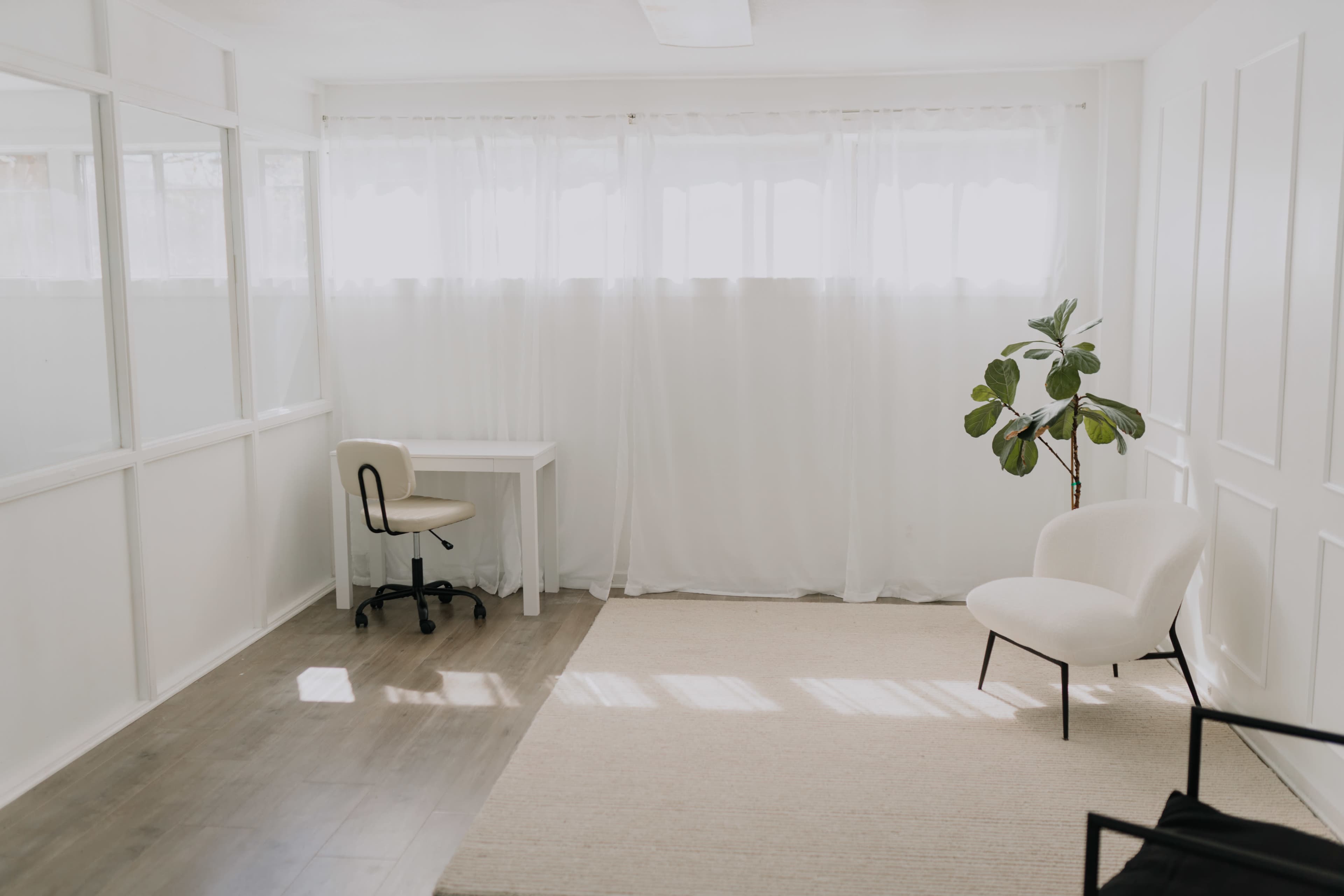 The image depicts a simple, bright room featuring a desk and chair, a potted plant, and large curtains allowing natural light to fill the space.