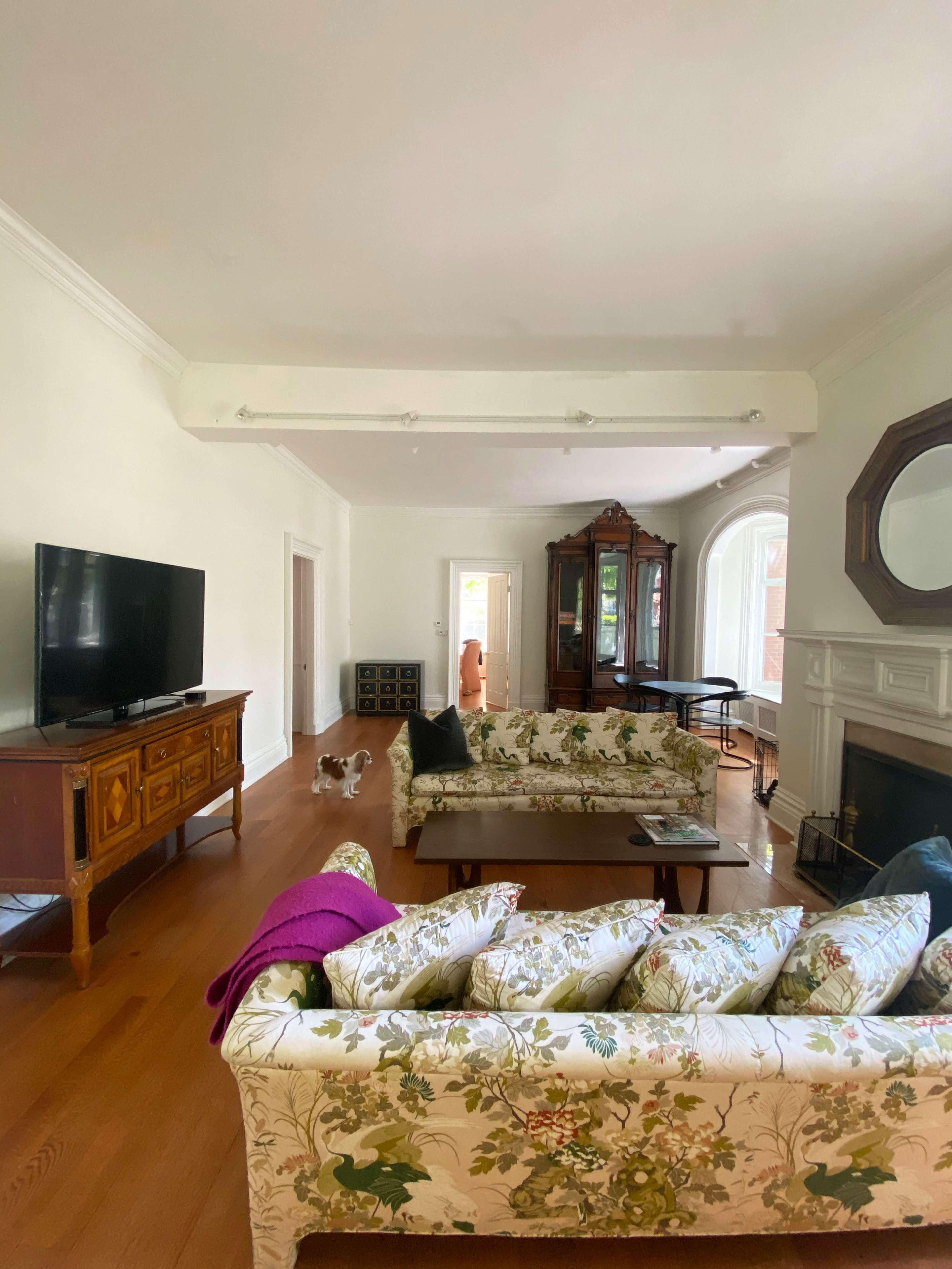 A spacious living room features two floral-patterned sofas facing a television, with a wooden cabinet and a door leading to another room in the background.