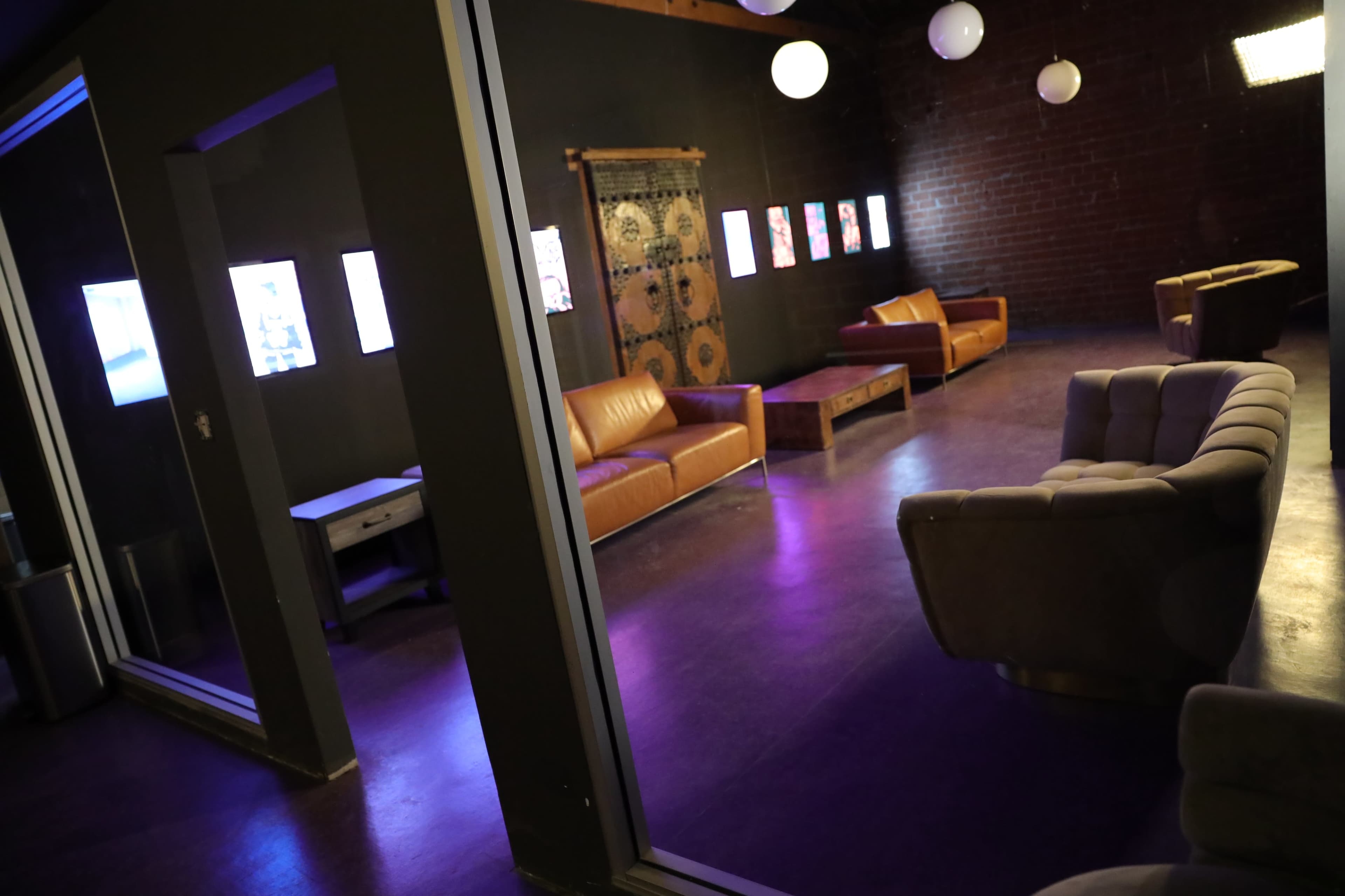 The image shows a dimly lit lounge area with brown leather couches, a coffee table, and wall art, surrounded by a dark-colored environment and illuminated by circular light fixtures.