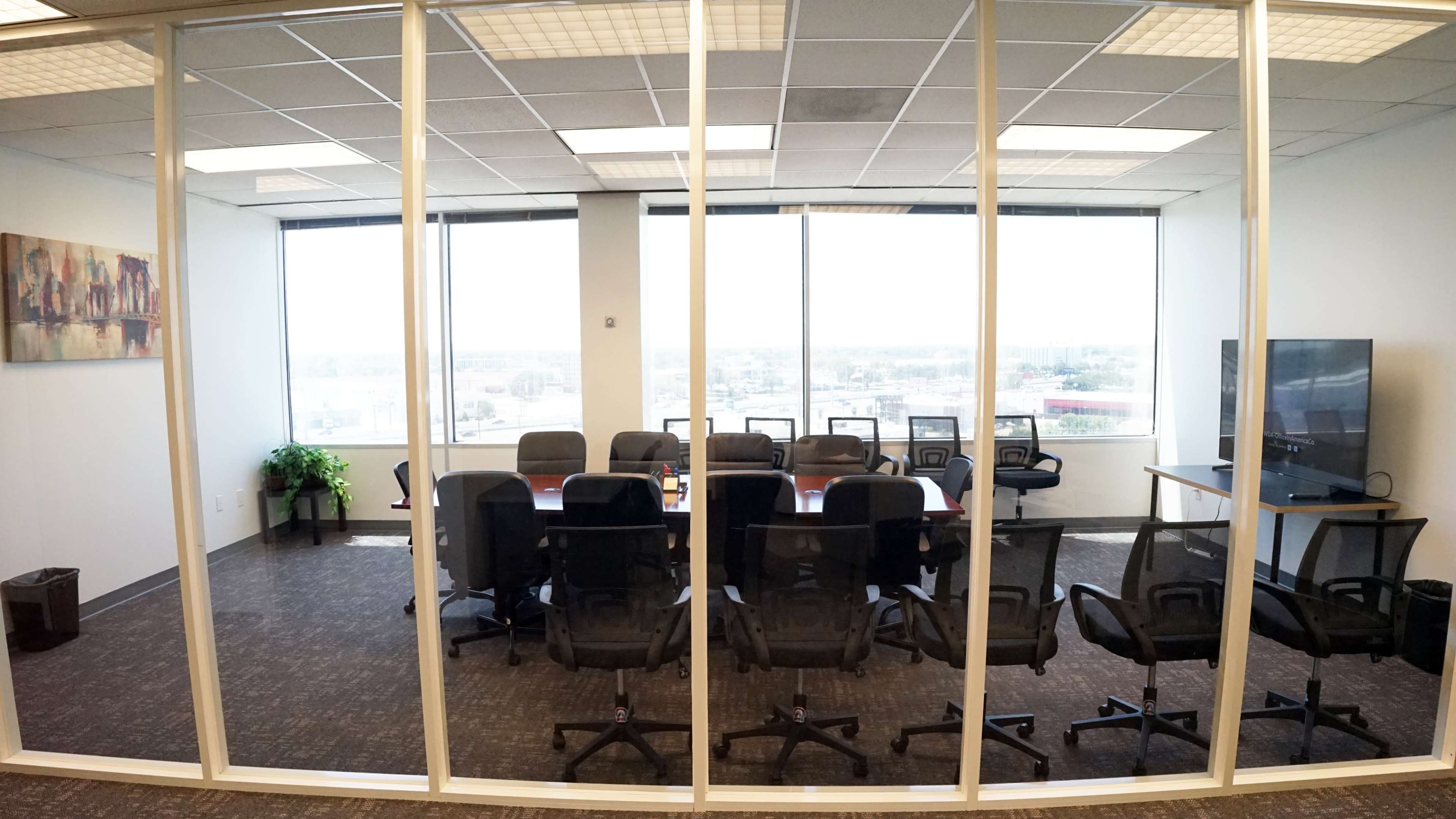 The image shows a glass-walled conference room furnished with a long table surrounded by several black chairs, with large windows offering an outside view.