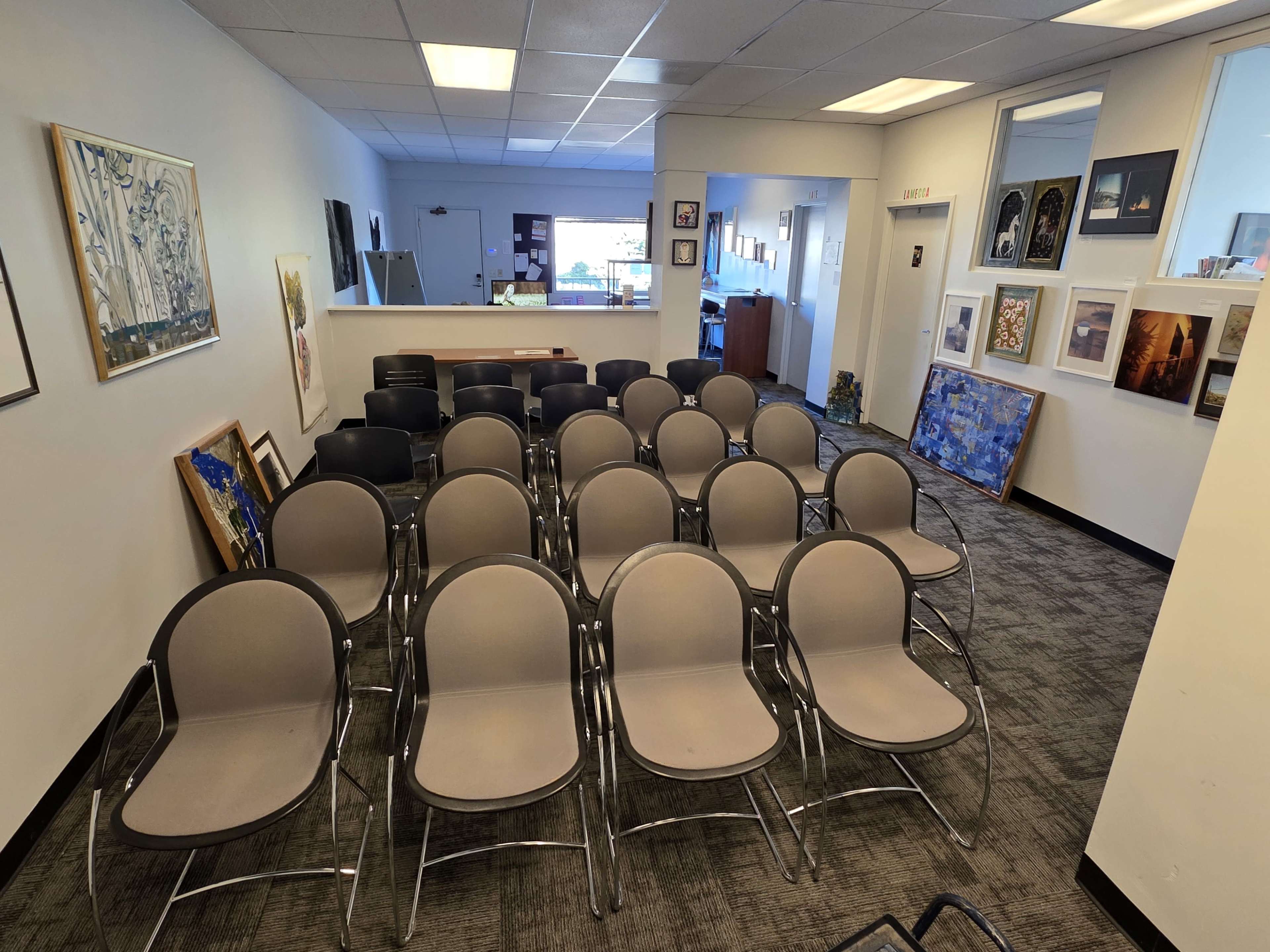 The image shows a series of gray chairs arranged in rows within a brightly lit office or gallery space, with artwork displayed on the walls.