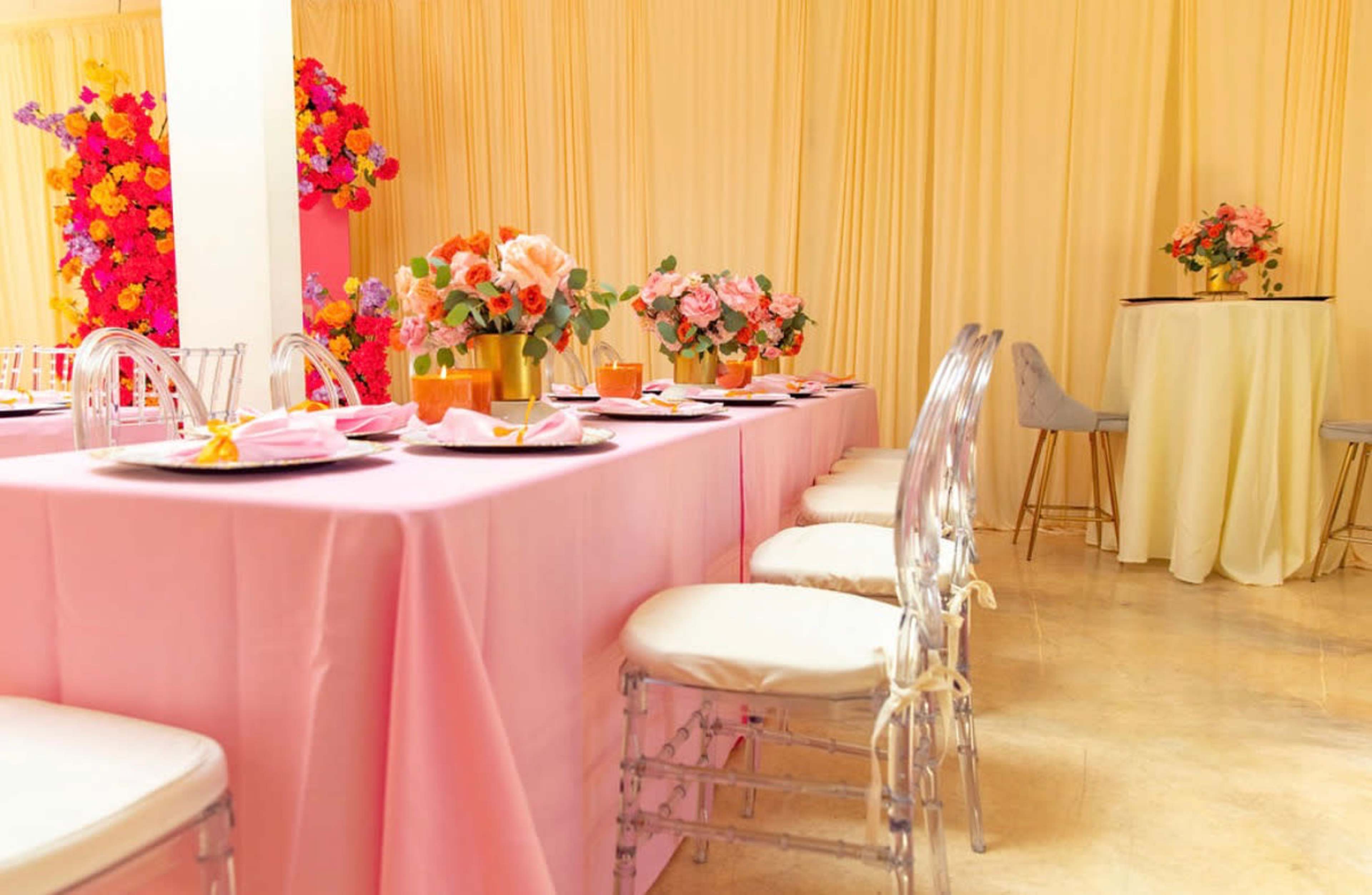 A long table dressed in pink linens is set with floral arrangements and clear chairs in a softly lit, elegant indoor space.